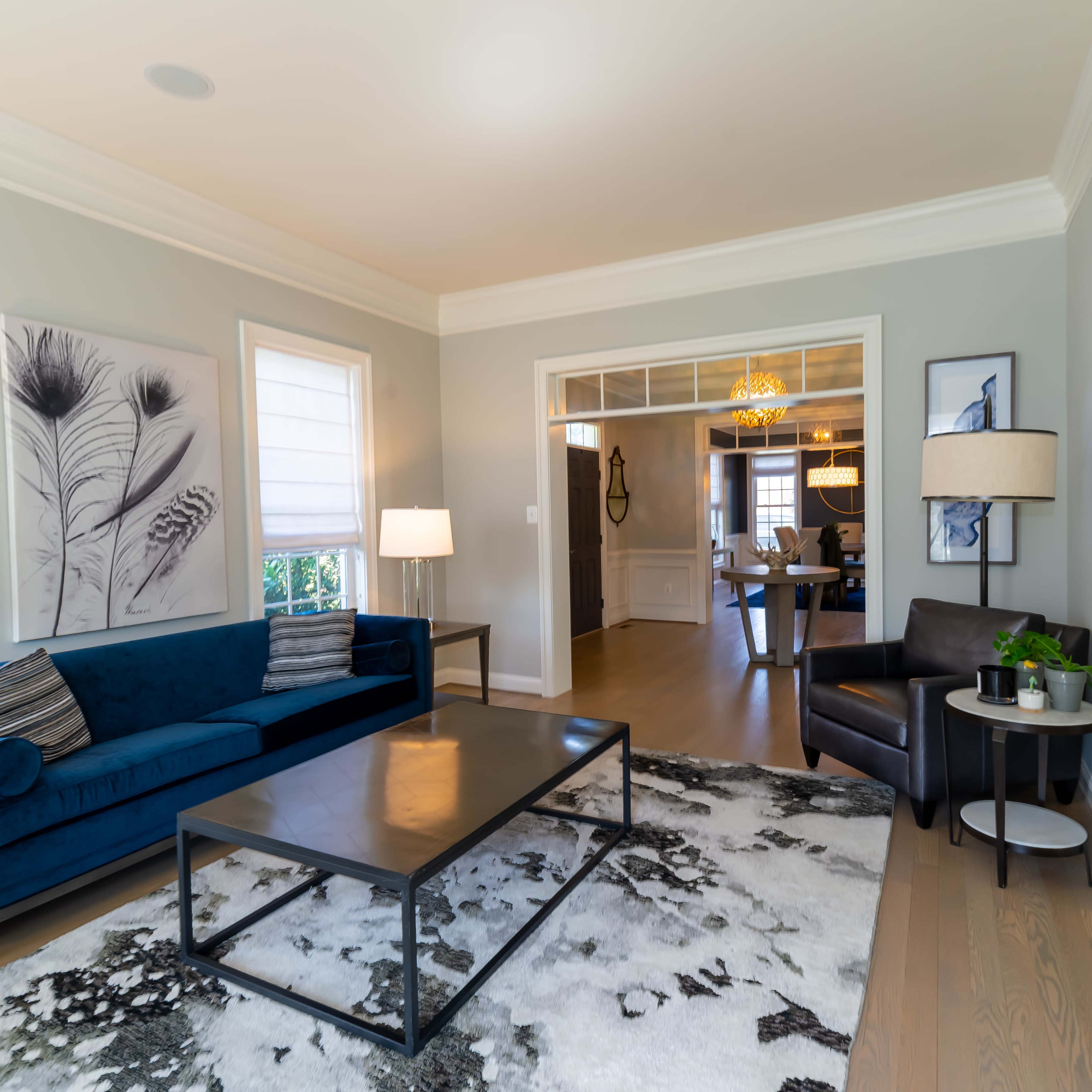 A modern living room features a blue sofa, a coffee table, and a black armchair, with artwork on the walls and a view into a dining area.