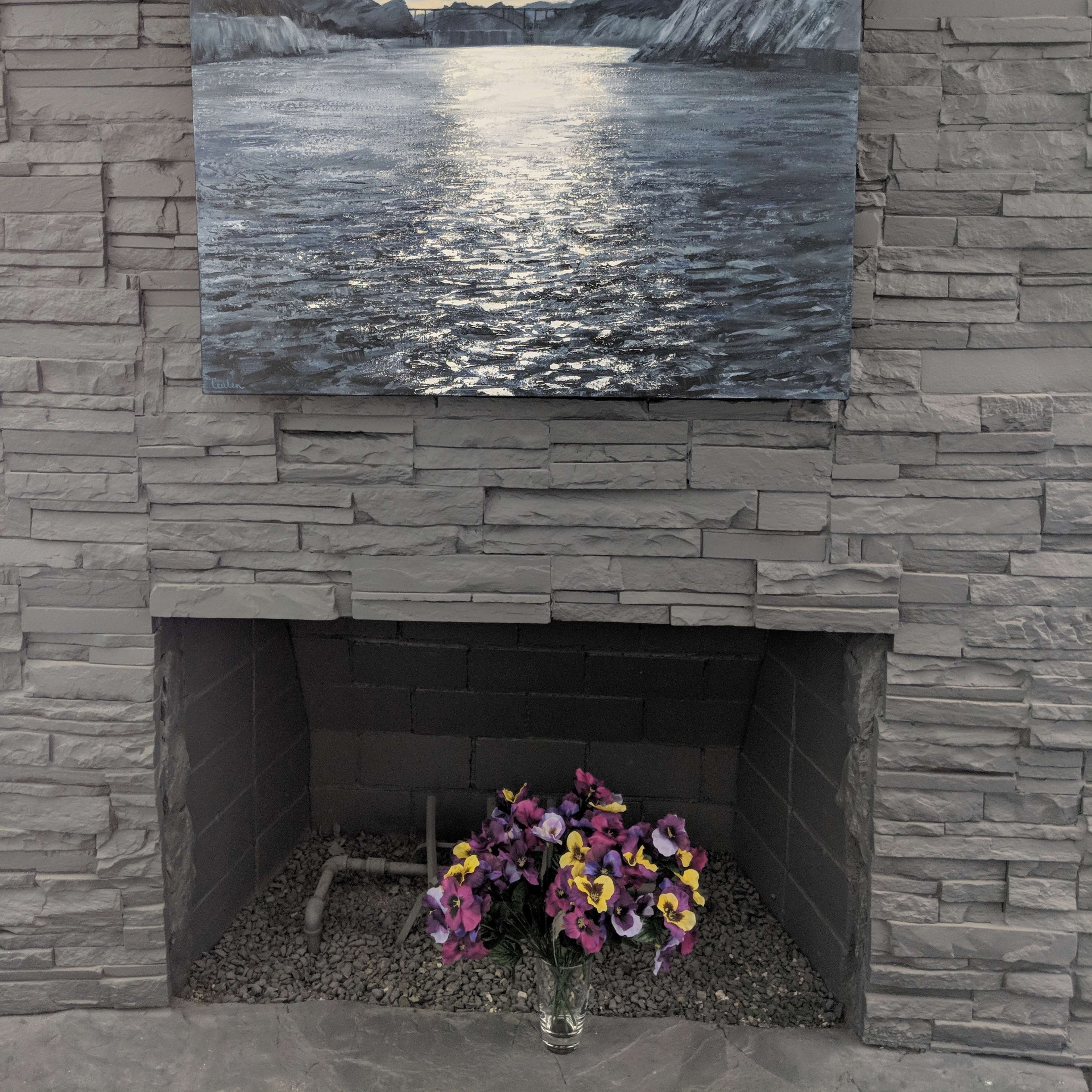 A large painting of a mountainous landscape is hung above a stone fireplace, with a vase of flowers in front.