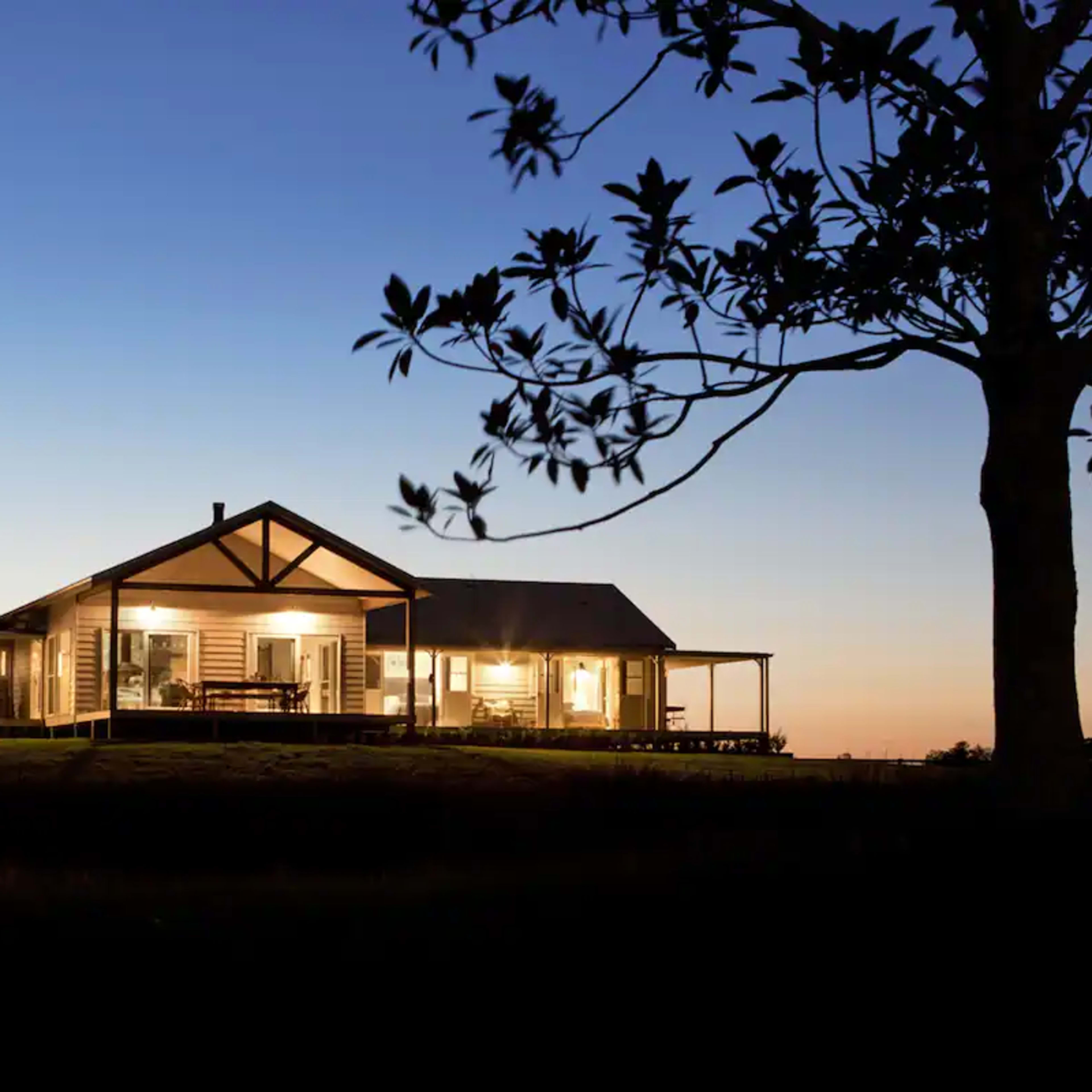 A large house with a veranda is illuminated at dusk, surrounded by trees and an open field.