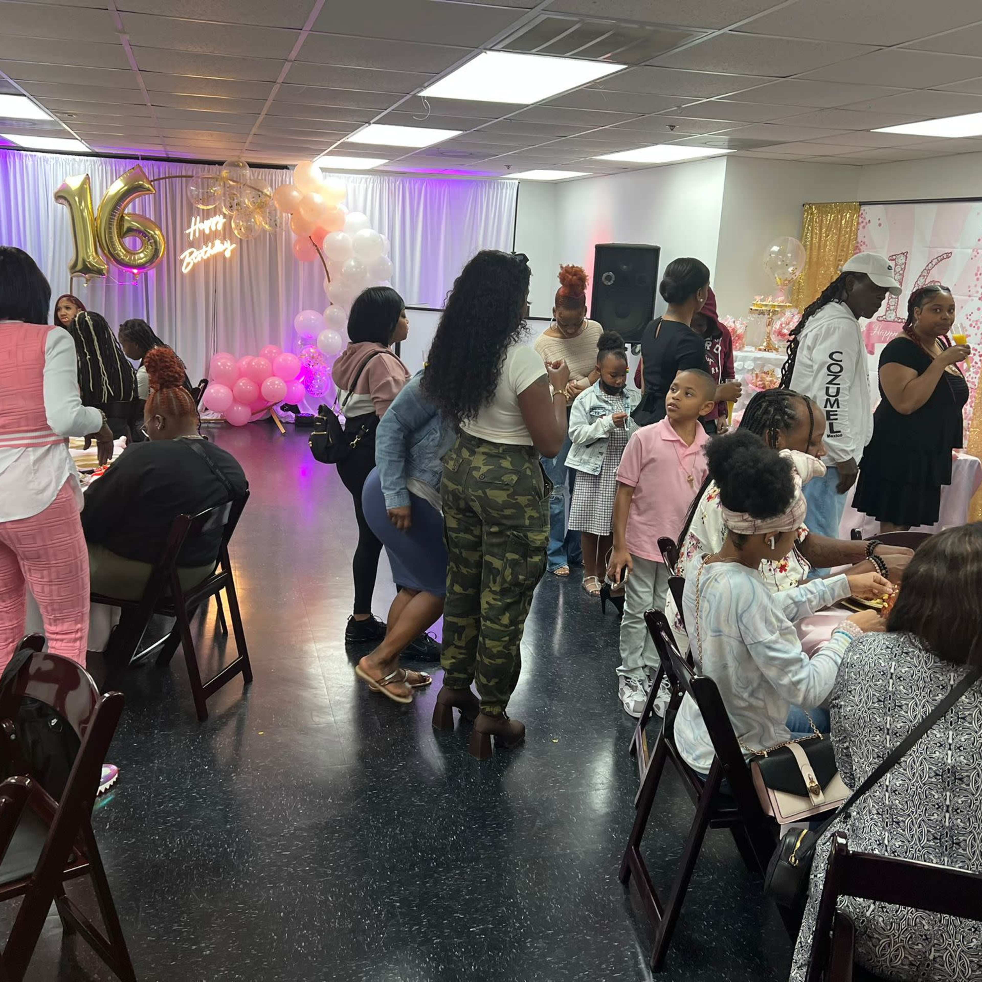 A gathering is taking place in a decorated room for a sweet sixteen celebration, with guests seated at tables, enjoying food and drinks while others socialize.