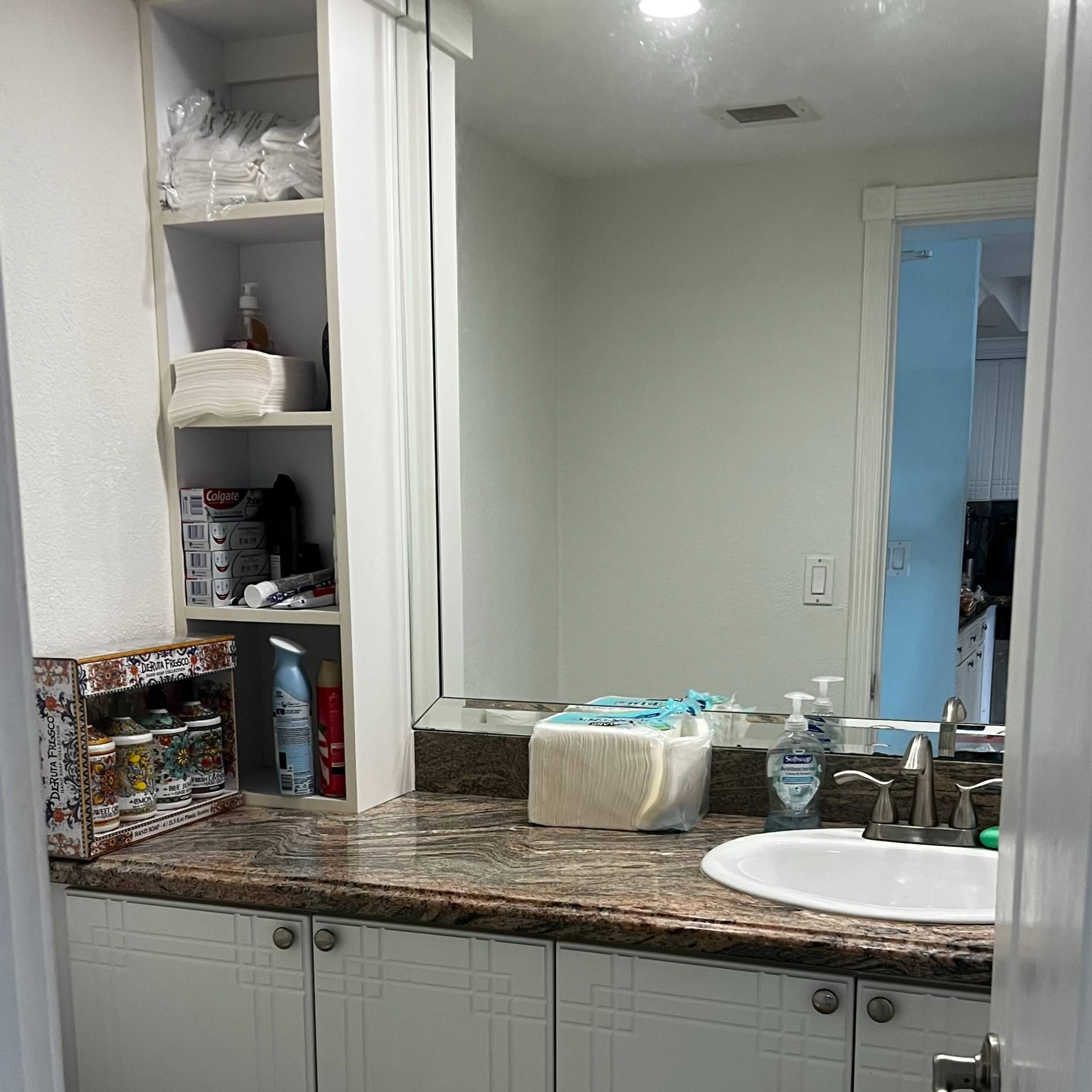 A bathroom with a white toilet, a sink with a mirror above, and shelves containing various toiletries and cleaning supplies.