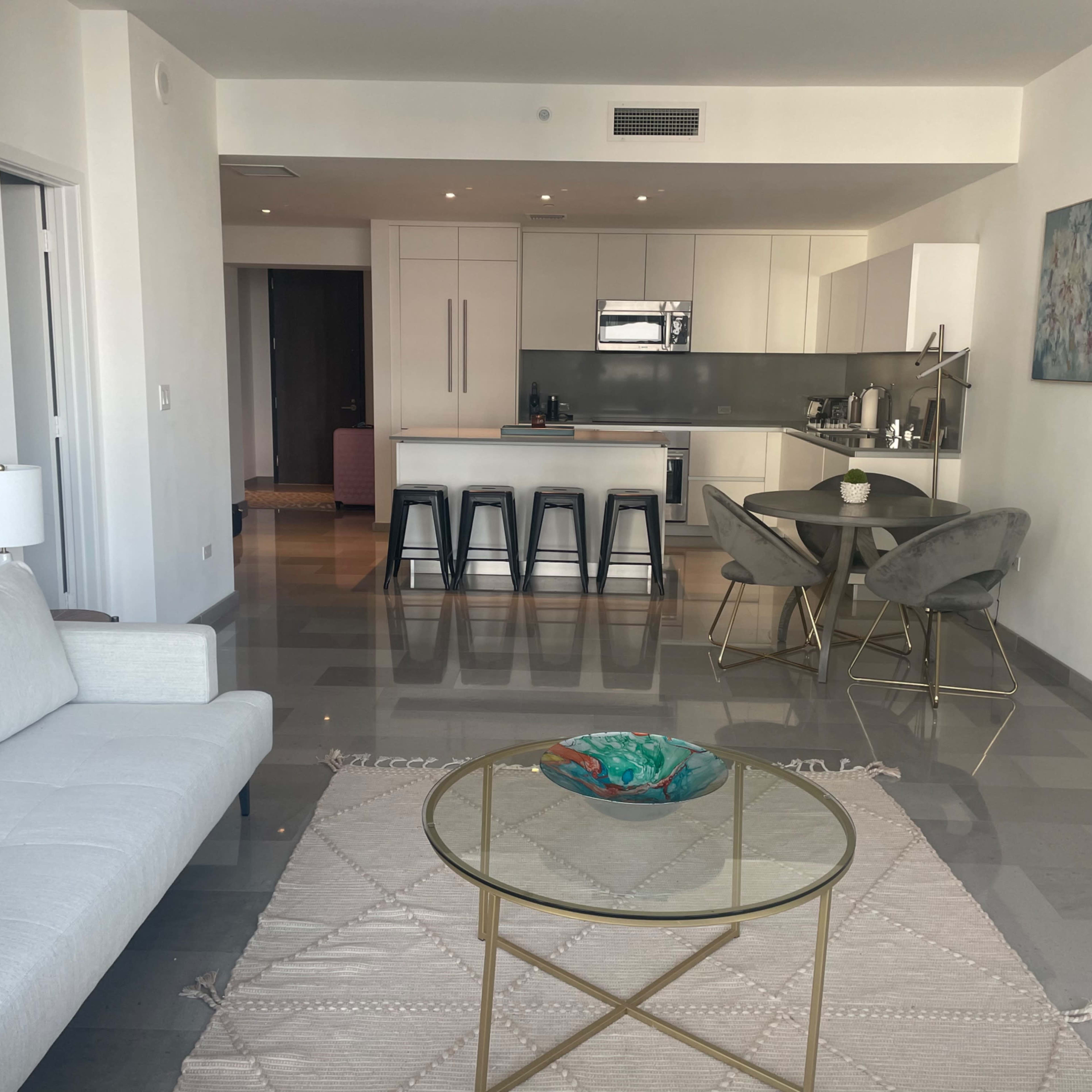 A modern living space featuring a white sofa, a round glass coffee table on a beige rug, and a kitchen area with bar stools and a dining table.