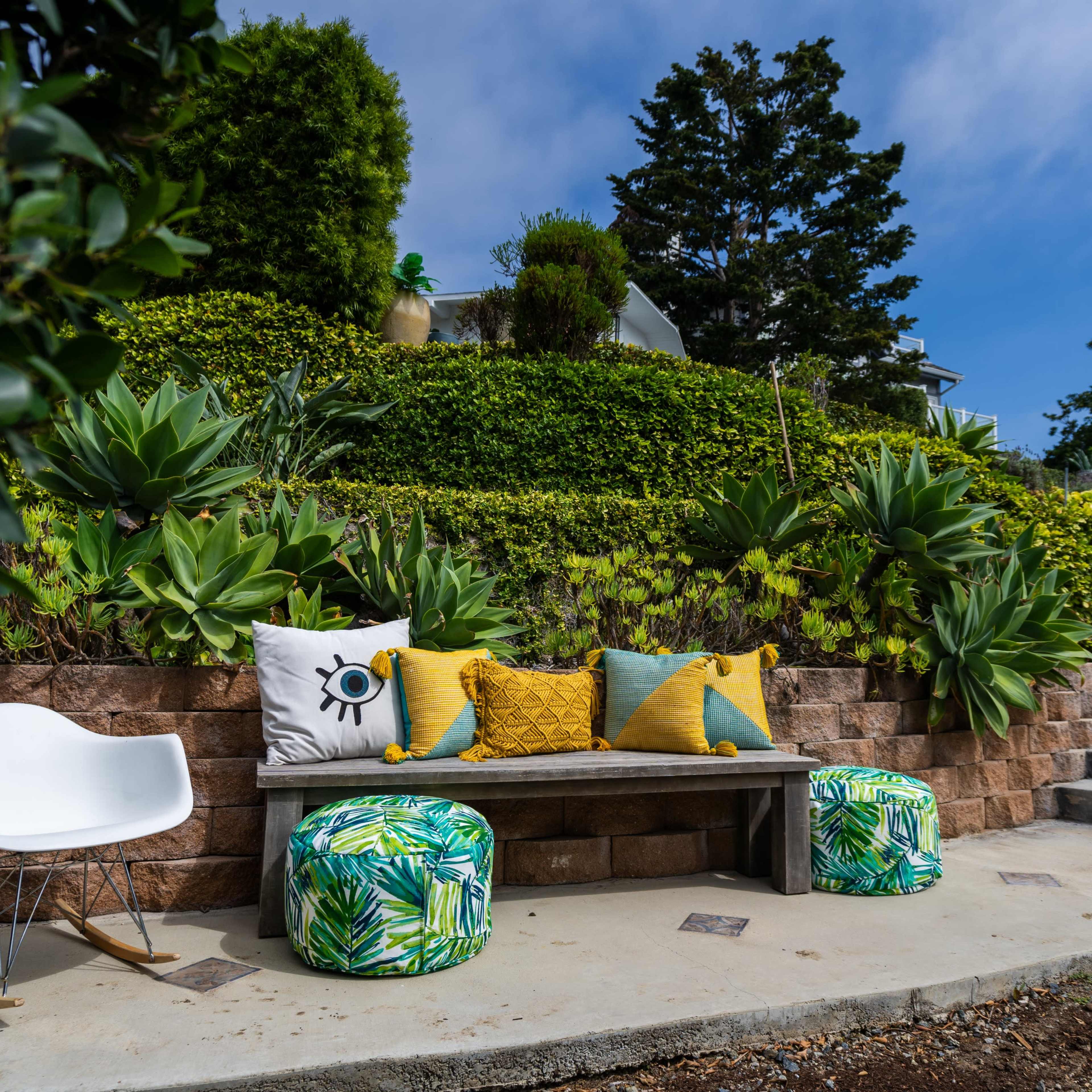 A stone bench with colorful cushions is situated in a landscaped garden surrounded by various plants and trees, leading up to a set of gray stairs.