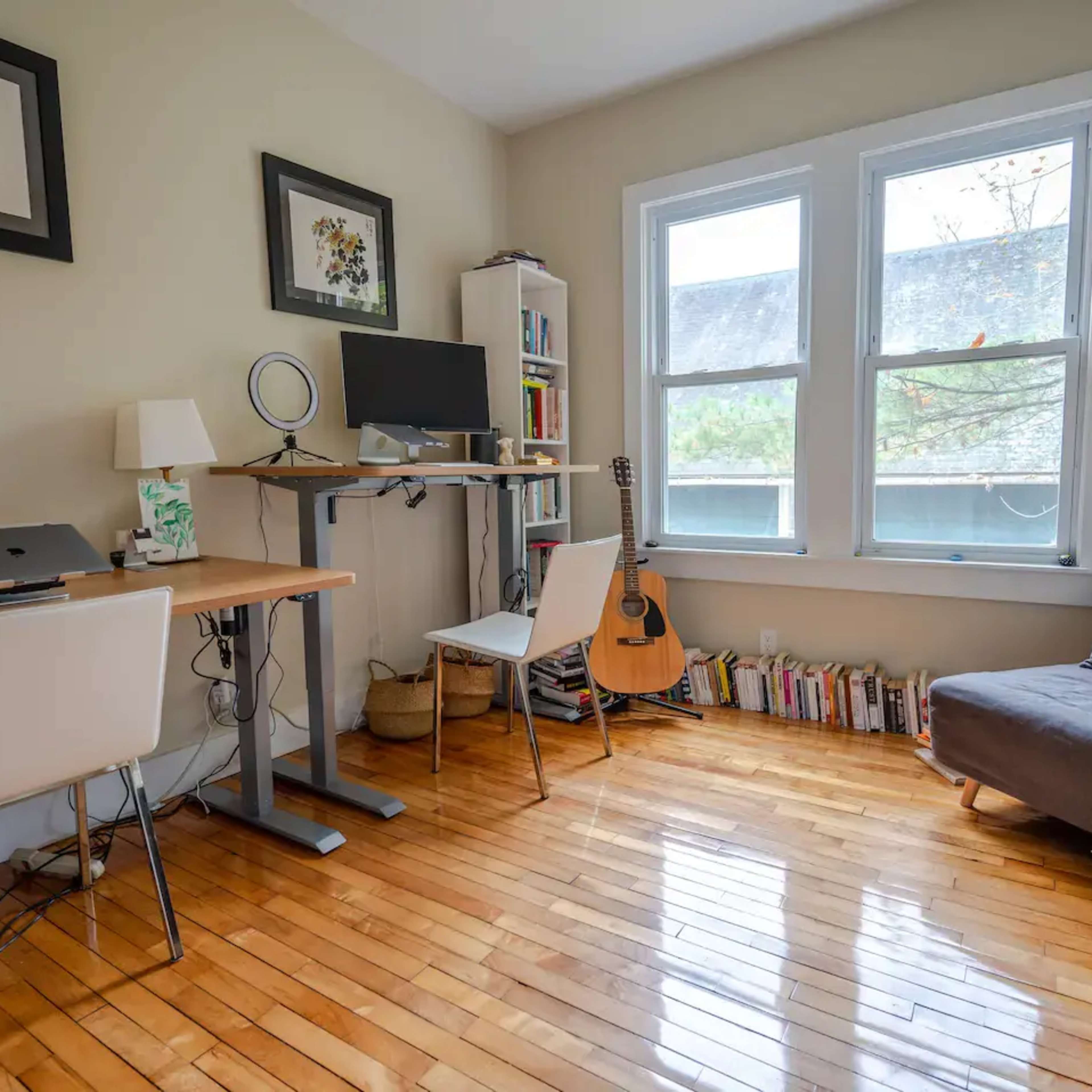 A room features a workspace with a desk and chair, a guitar resting against the wall, a small bookshelf, and large windows allowing natural light.