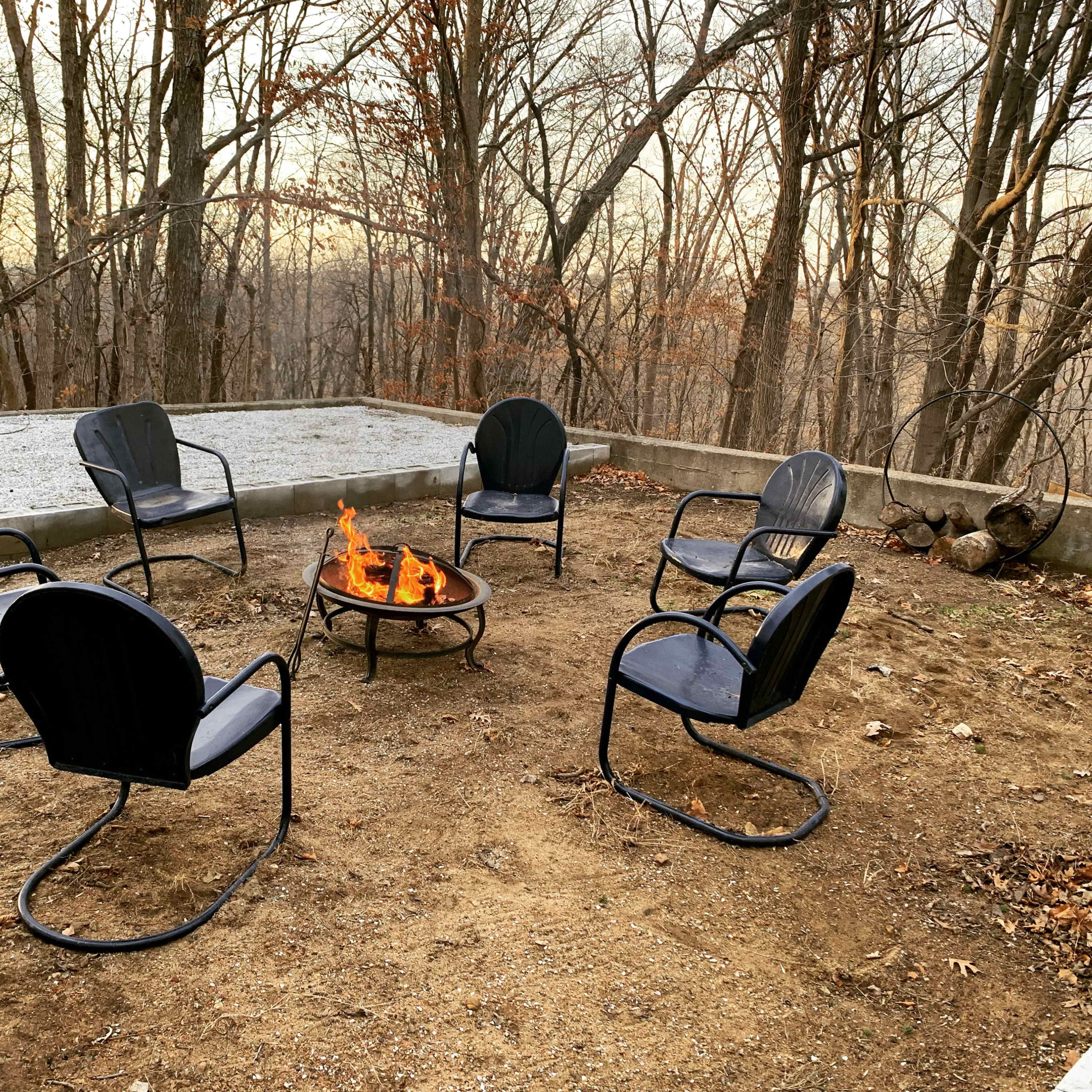 A circular arrangement of six black chairs surrounds a fire pit in a wooded area during dusk.