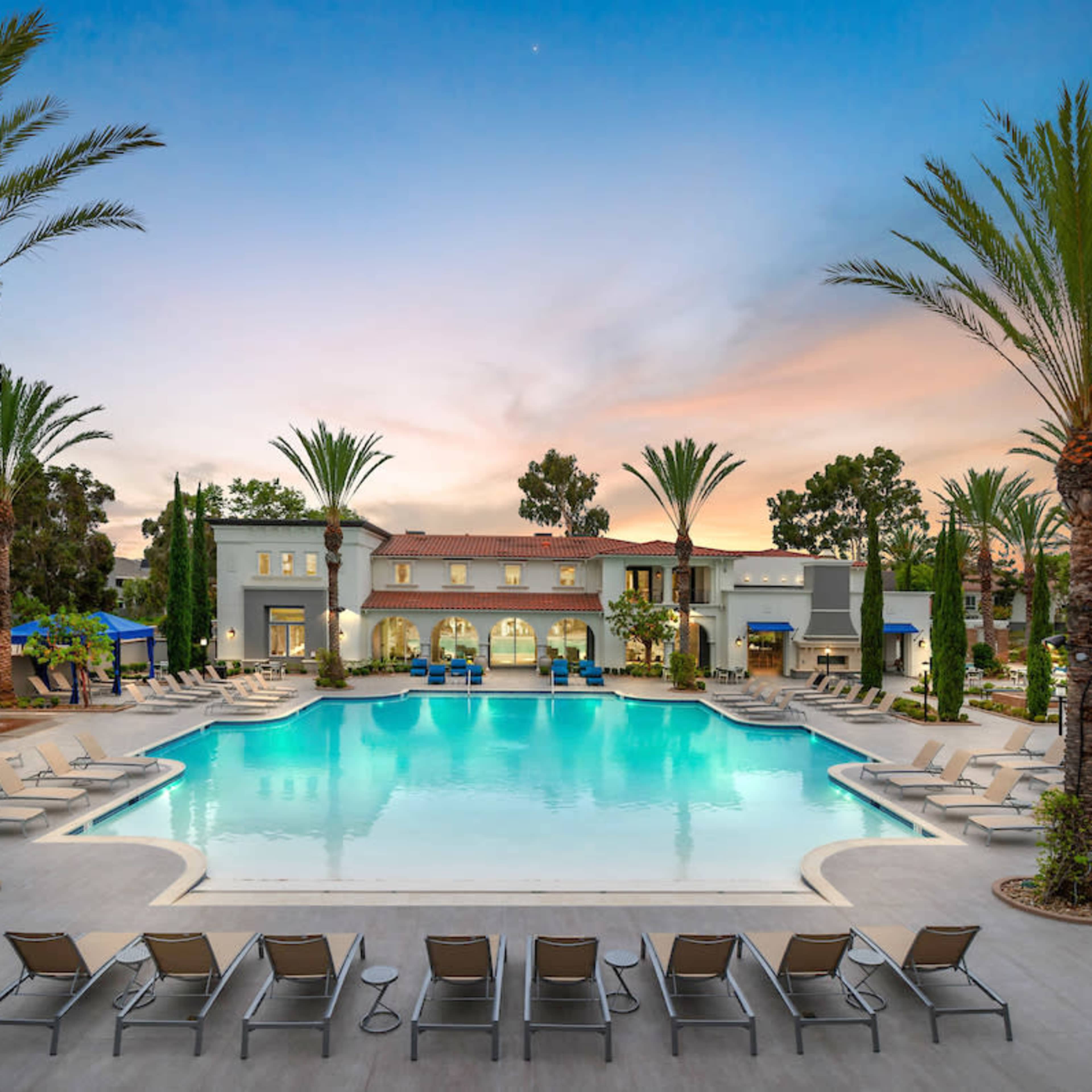 A large swimming pool surrounded by palm trees and lounge chairs, with a building in the background under a colorful sky.