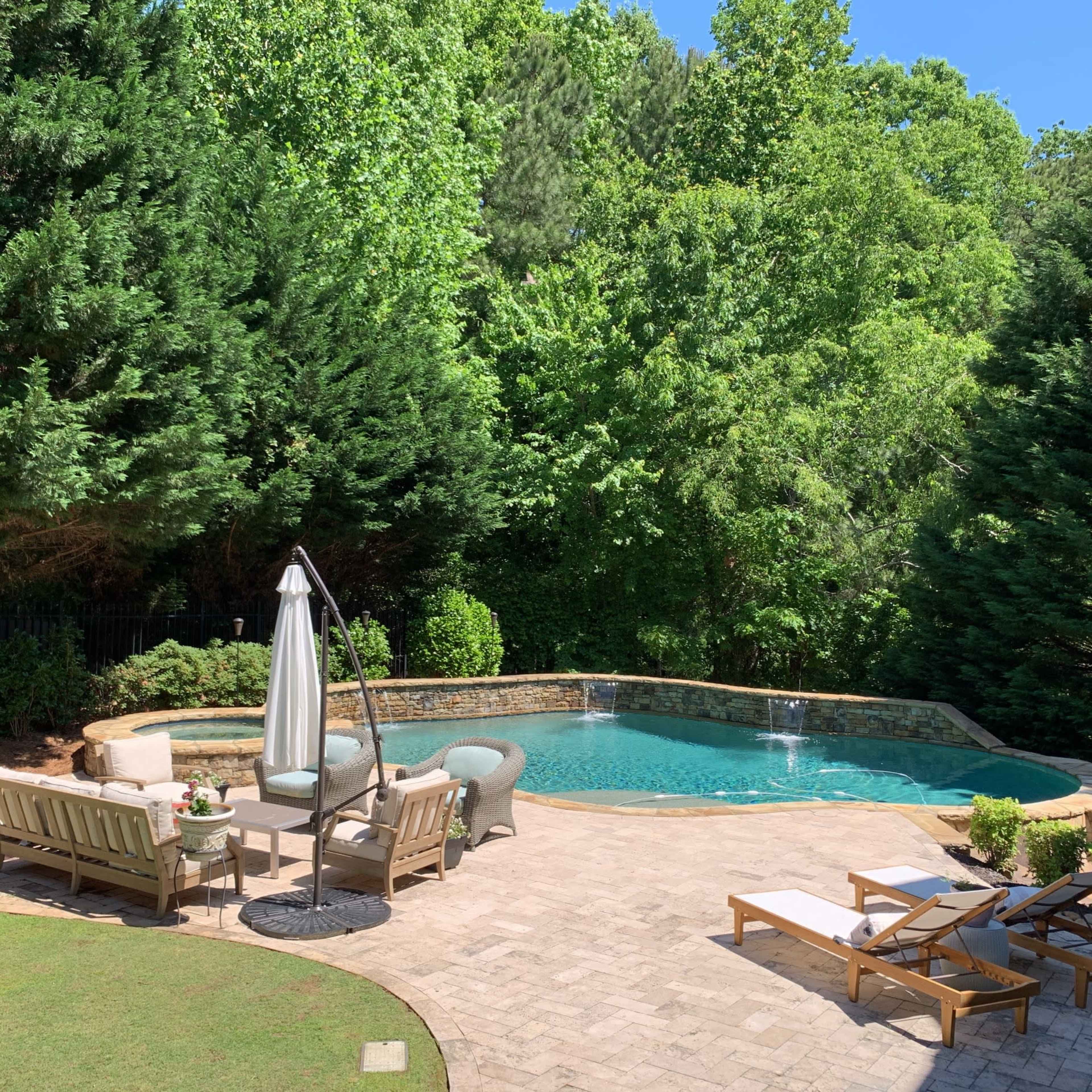 A landscaped backyard features a swimming pool surrounded by patio chairs and tables under a blue sky.