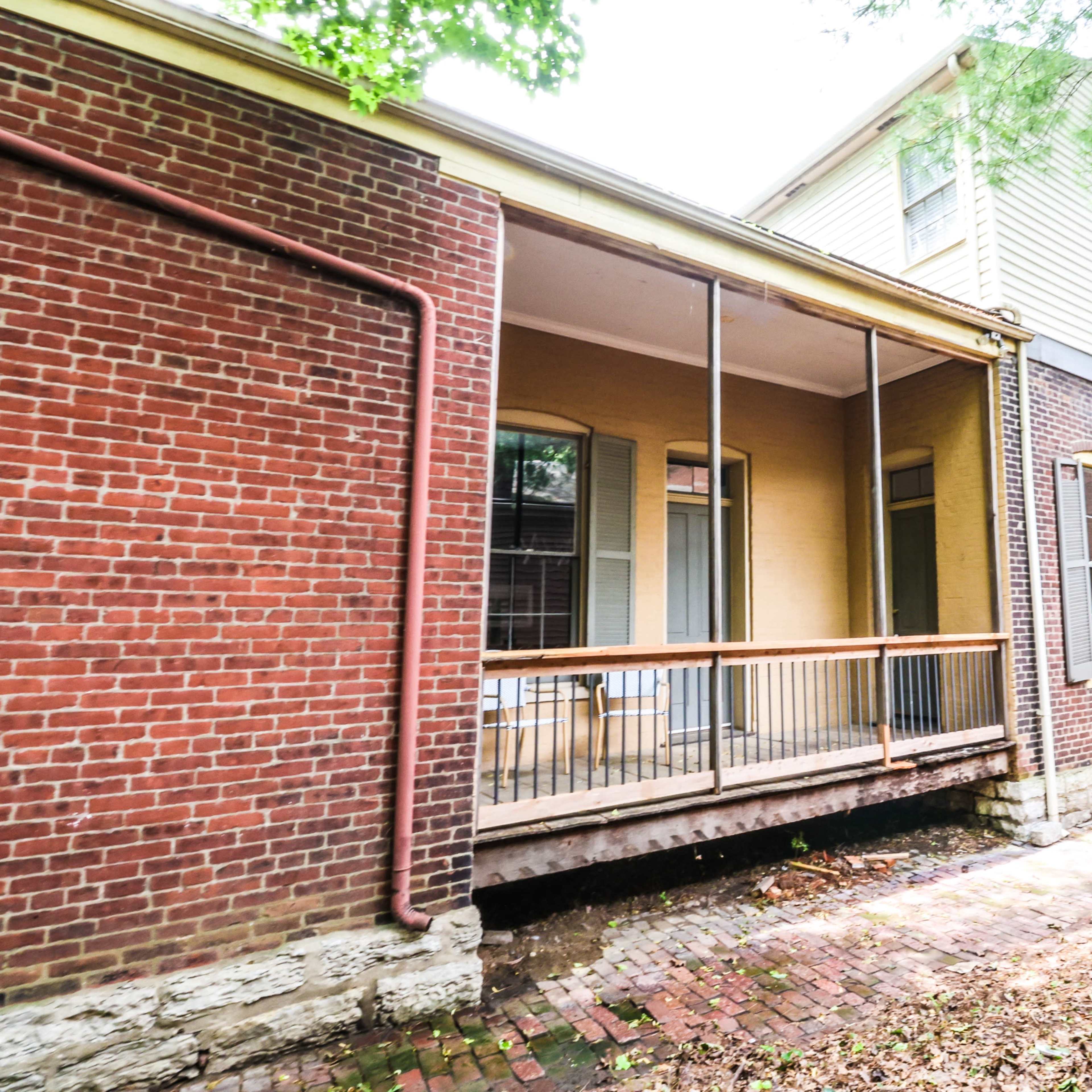 A brick house with a porch covered by a railing, surrounded by trees and a pathway made of bricks.