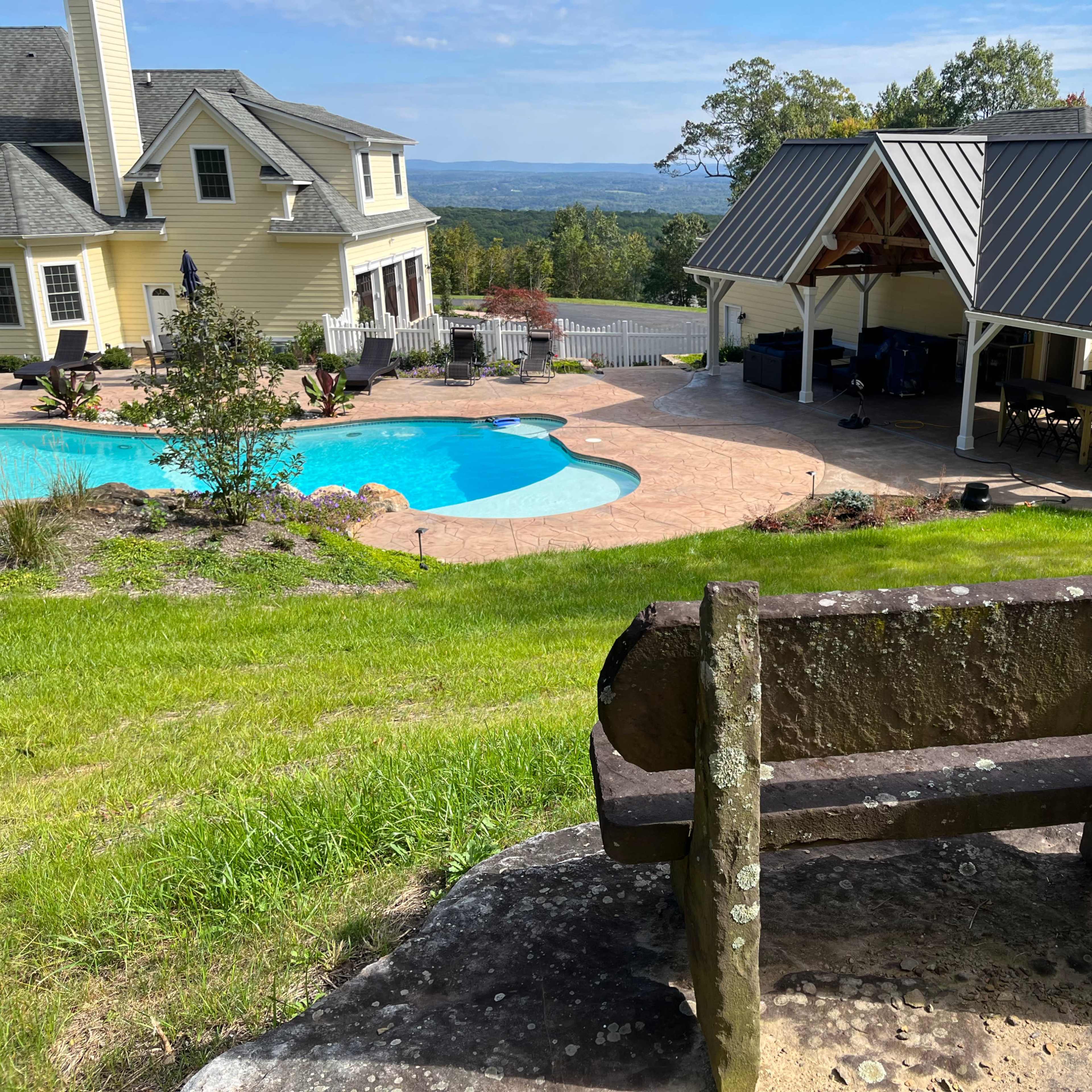A wooden bench overlooks a landscaped backyard with a swimming pool and a covered patio area beside a house.