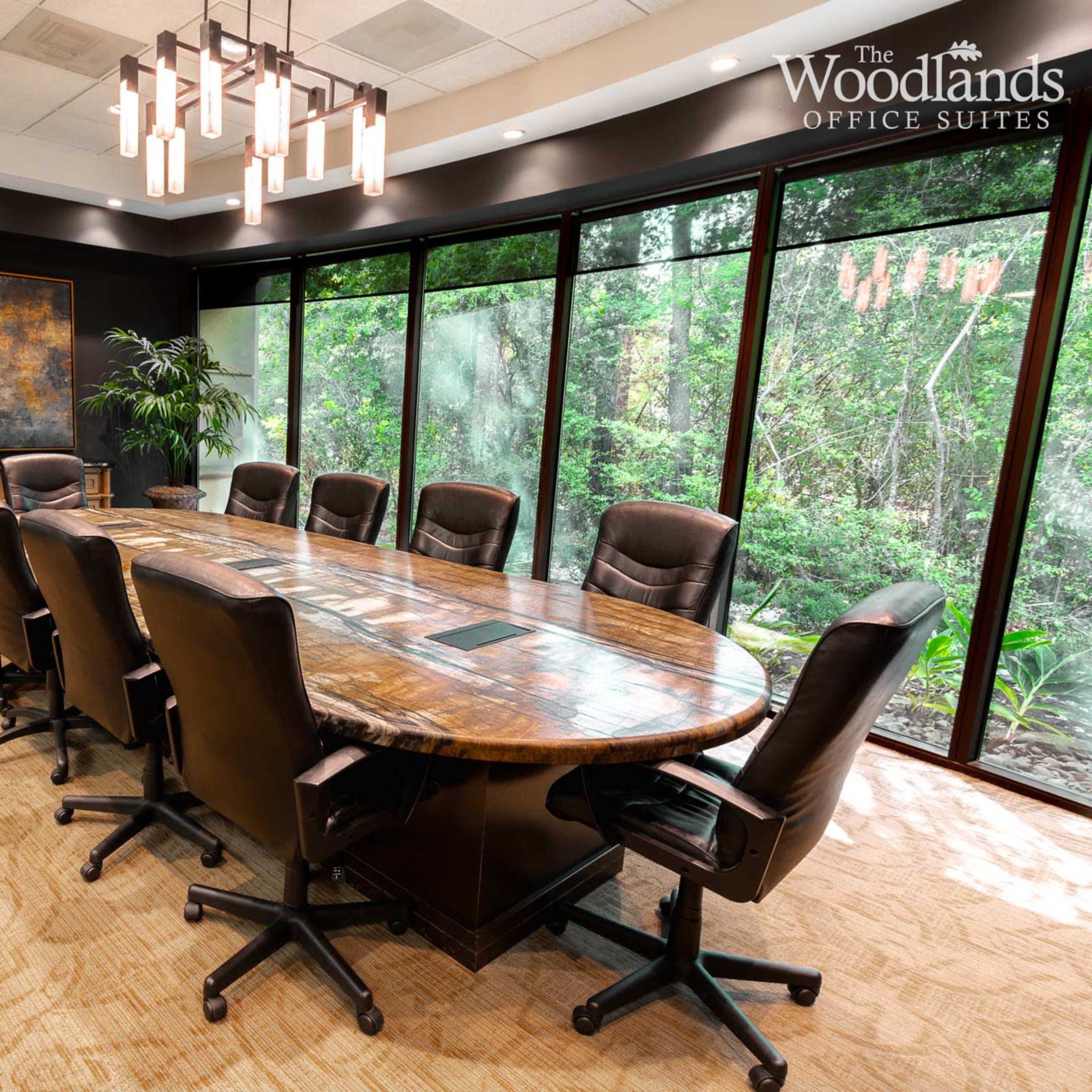A conference room features a large round wooden table surrounded by executive chairs, with large windows overlooking a green landscape.
