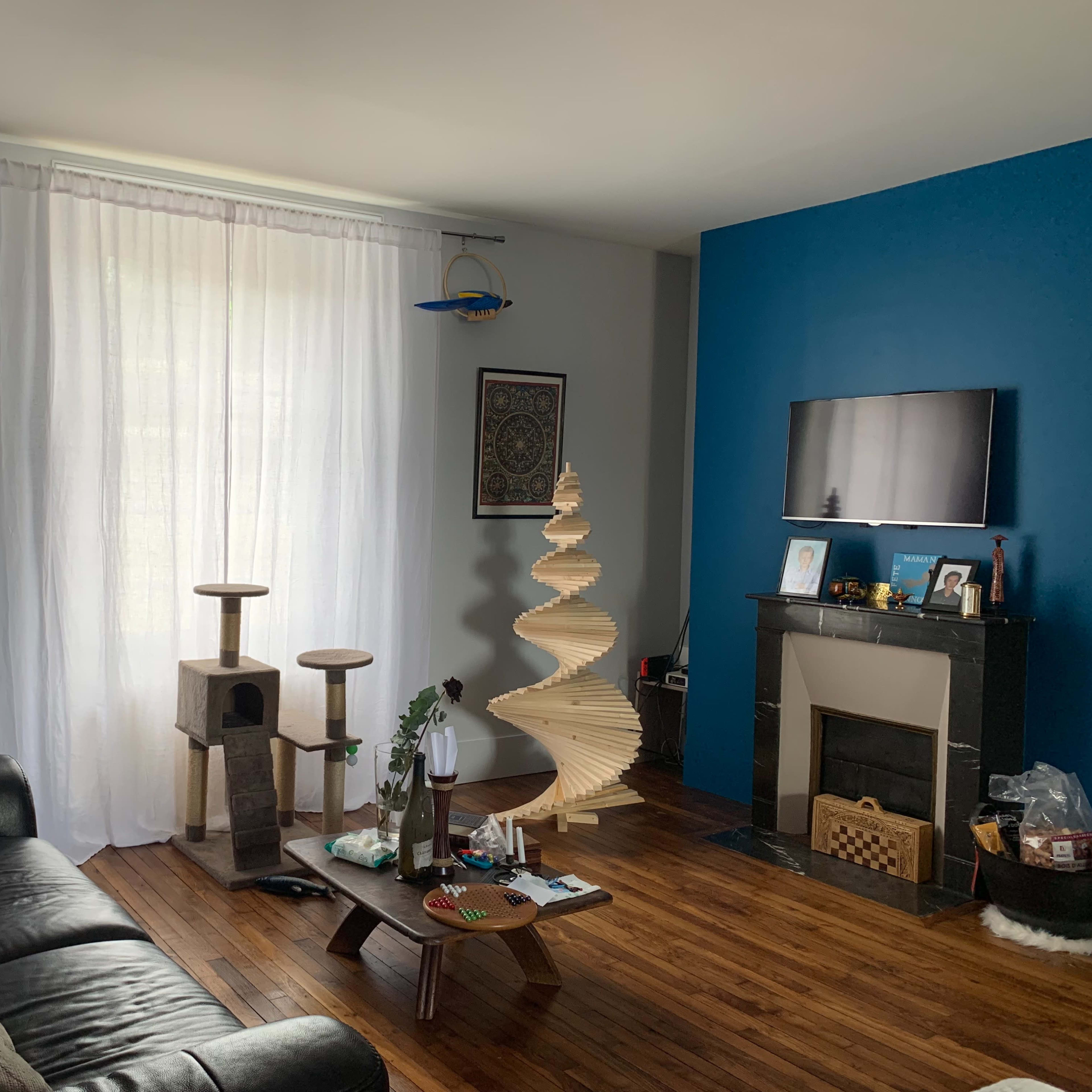 A living room with a black leather sofa, a wooden floor, a tall decorative cat tree, a spiral bookshelf, and a wall-mounted TV against a blue feature wall.
