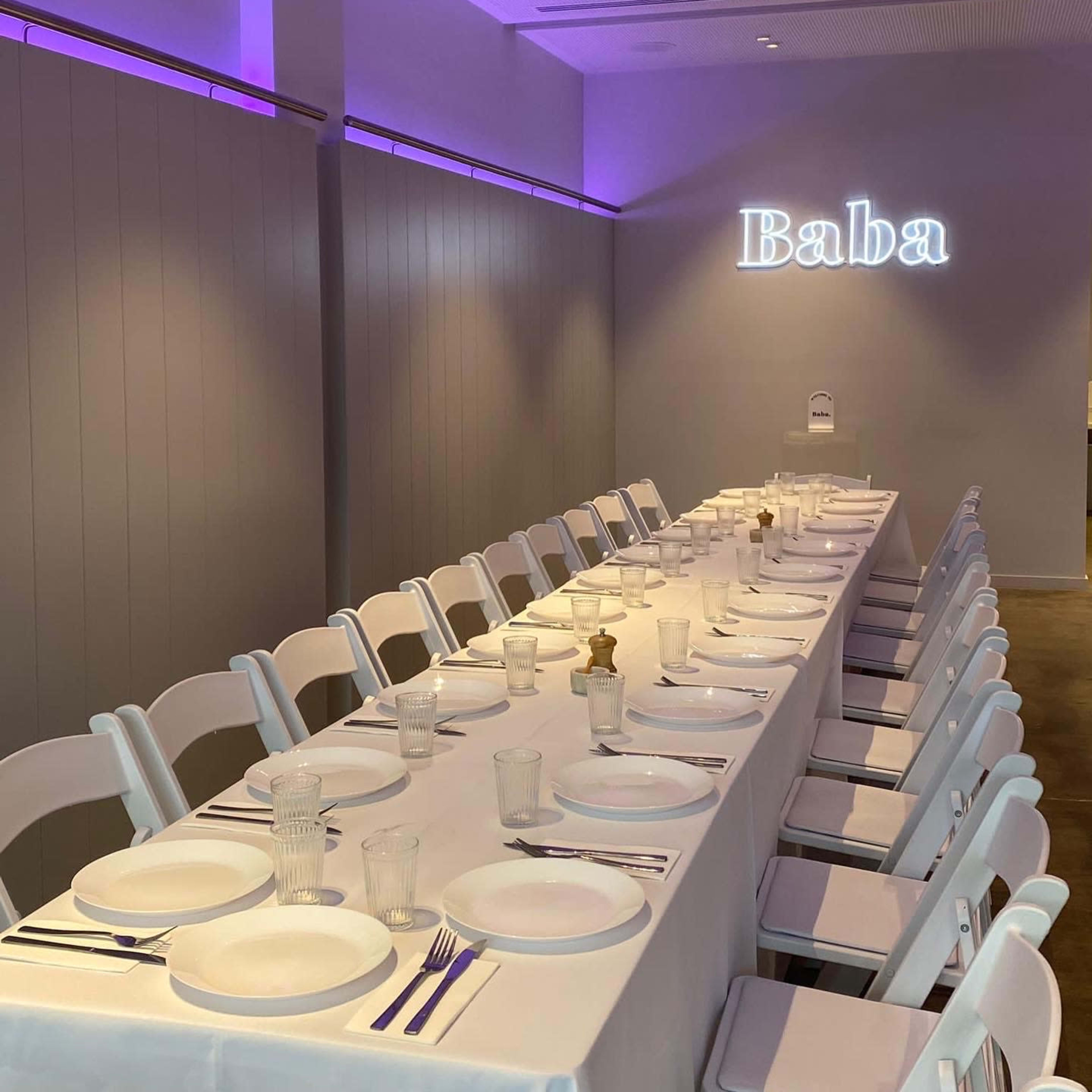 A long, neatly set dining table with white tableware and clear glasses is illuminated by purple lighting, with the word "Baba" displayed on the wall.
