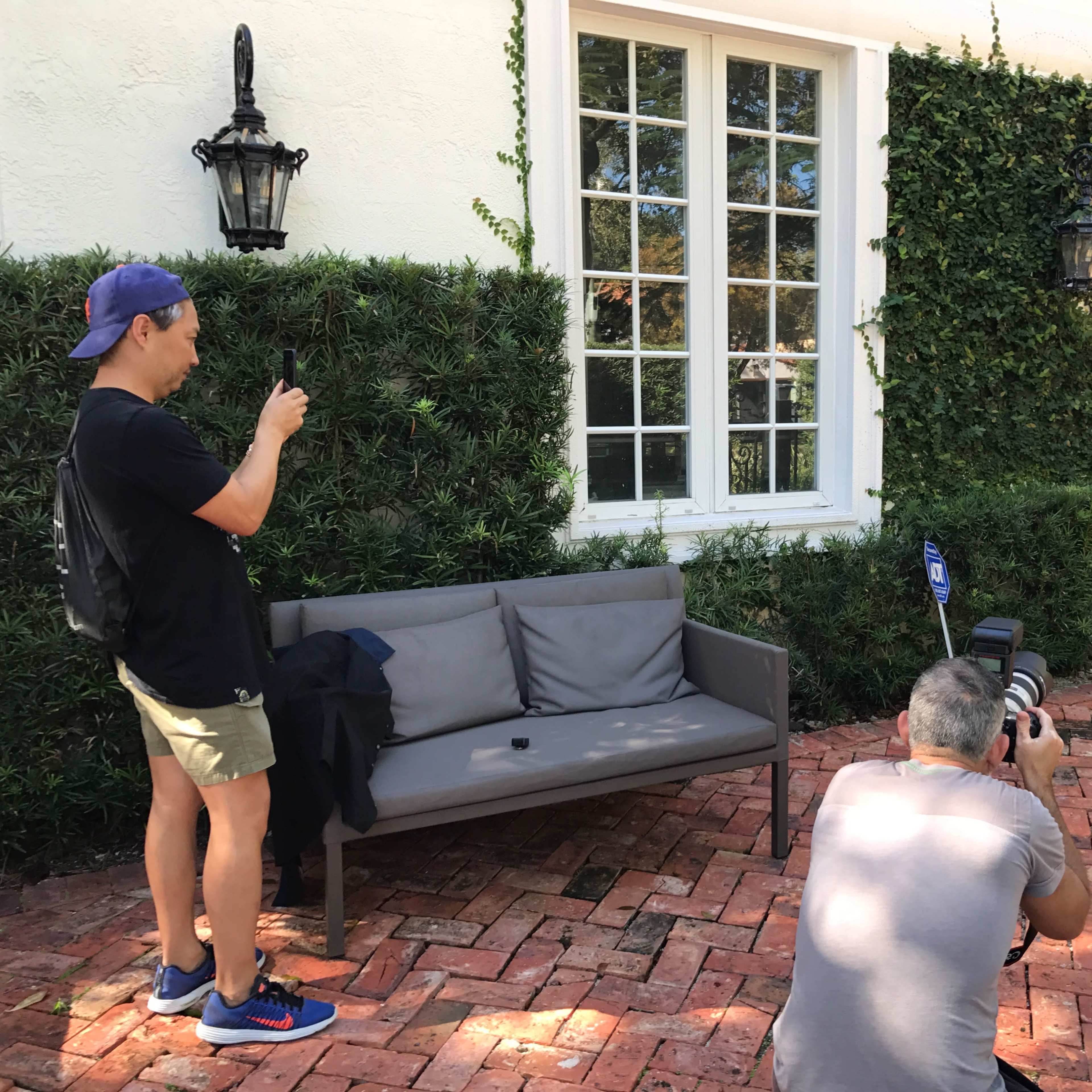 A photographer captures a male model posing in swimwear outside a well-lit home, while another person takes photos with a smartphone nearby.