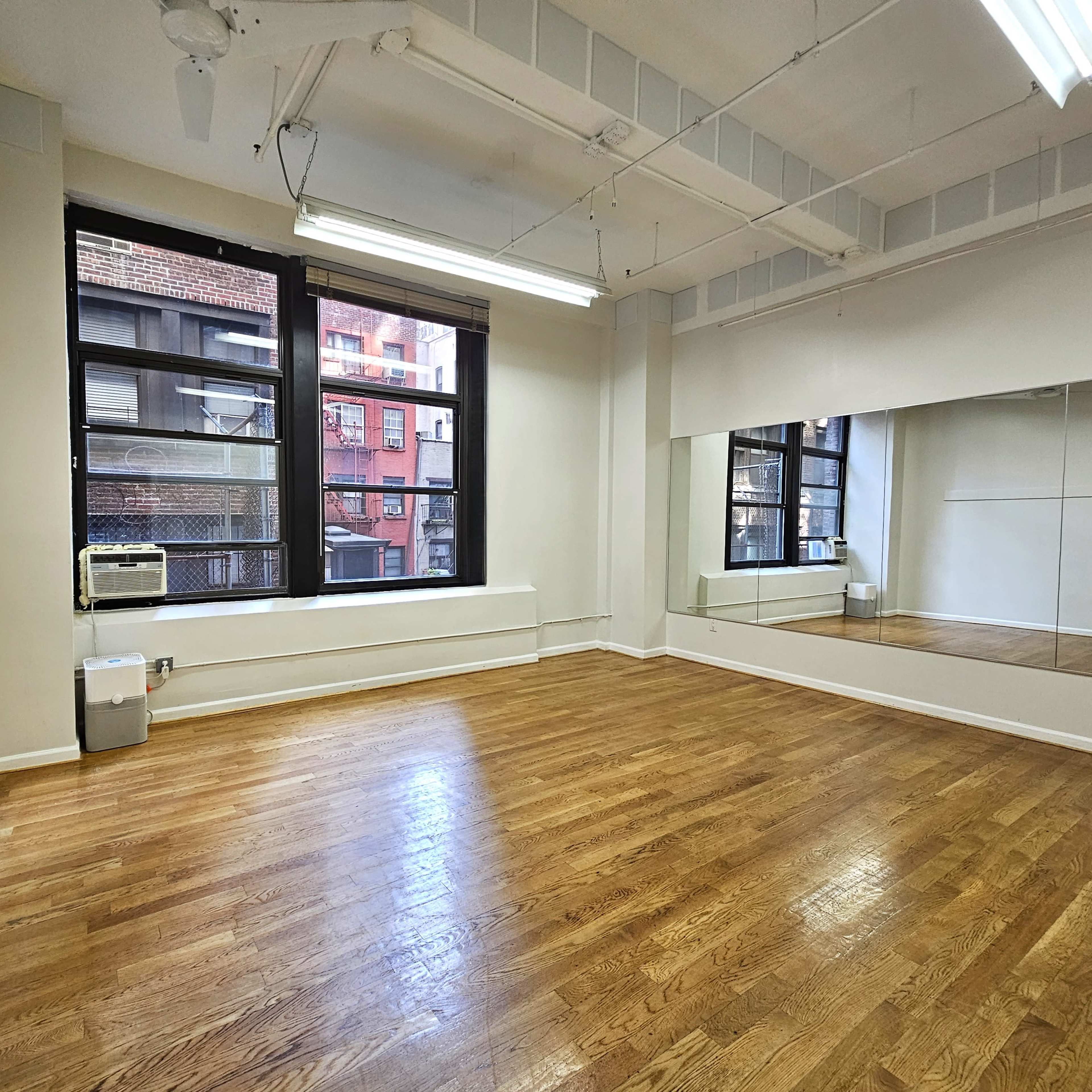 A spacious, empty room with wooden floors, large windows, and a mirror along one wall.