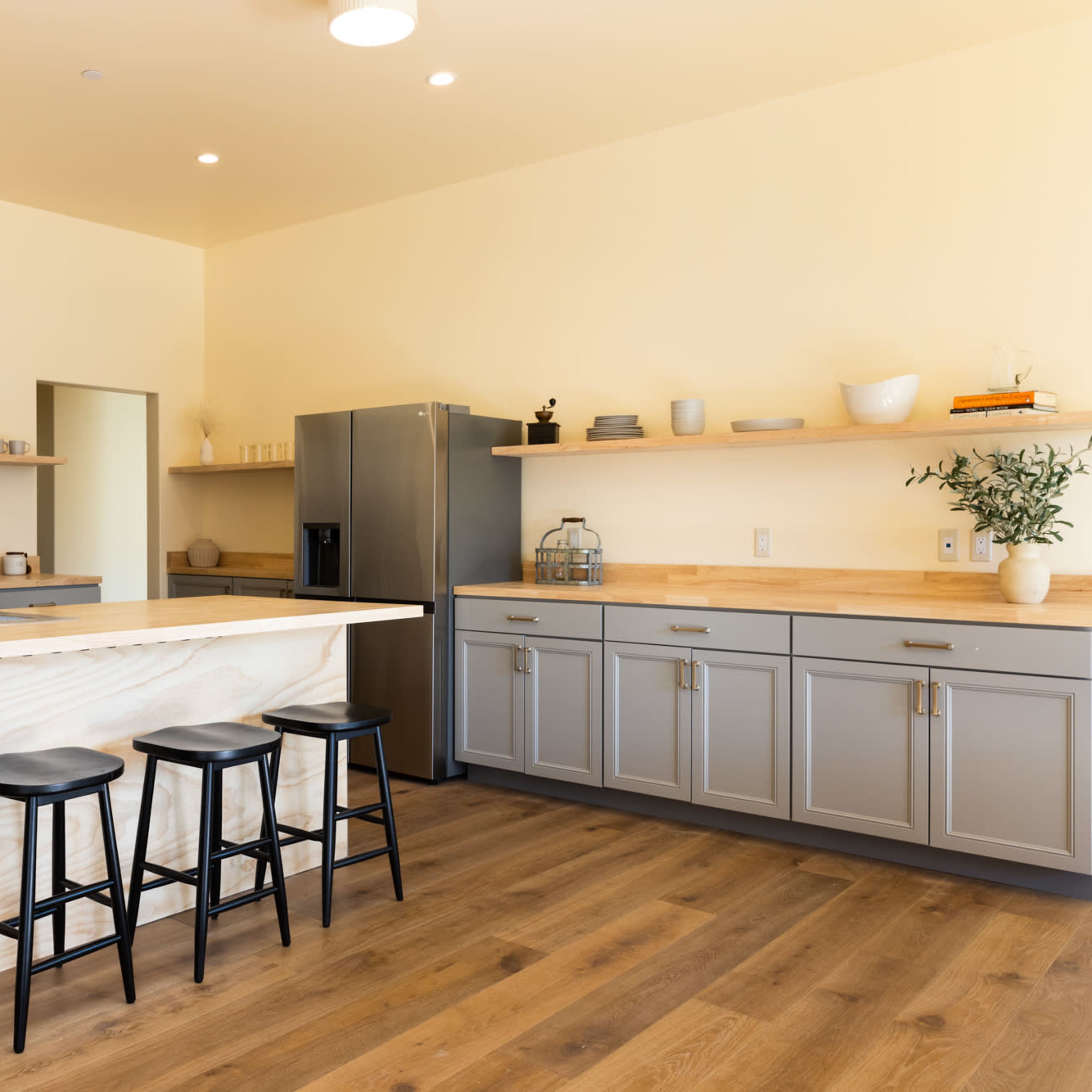 A modern kitchen featuring a central island with three black stools, stainless steel appliances, and light-colored cabinetry against wooden flooring.