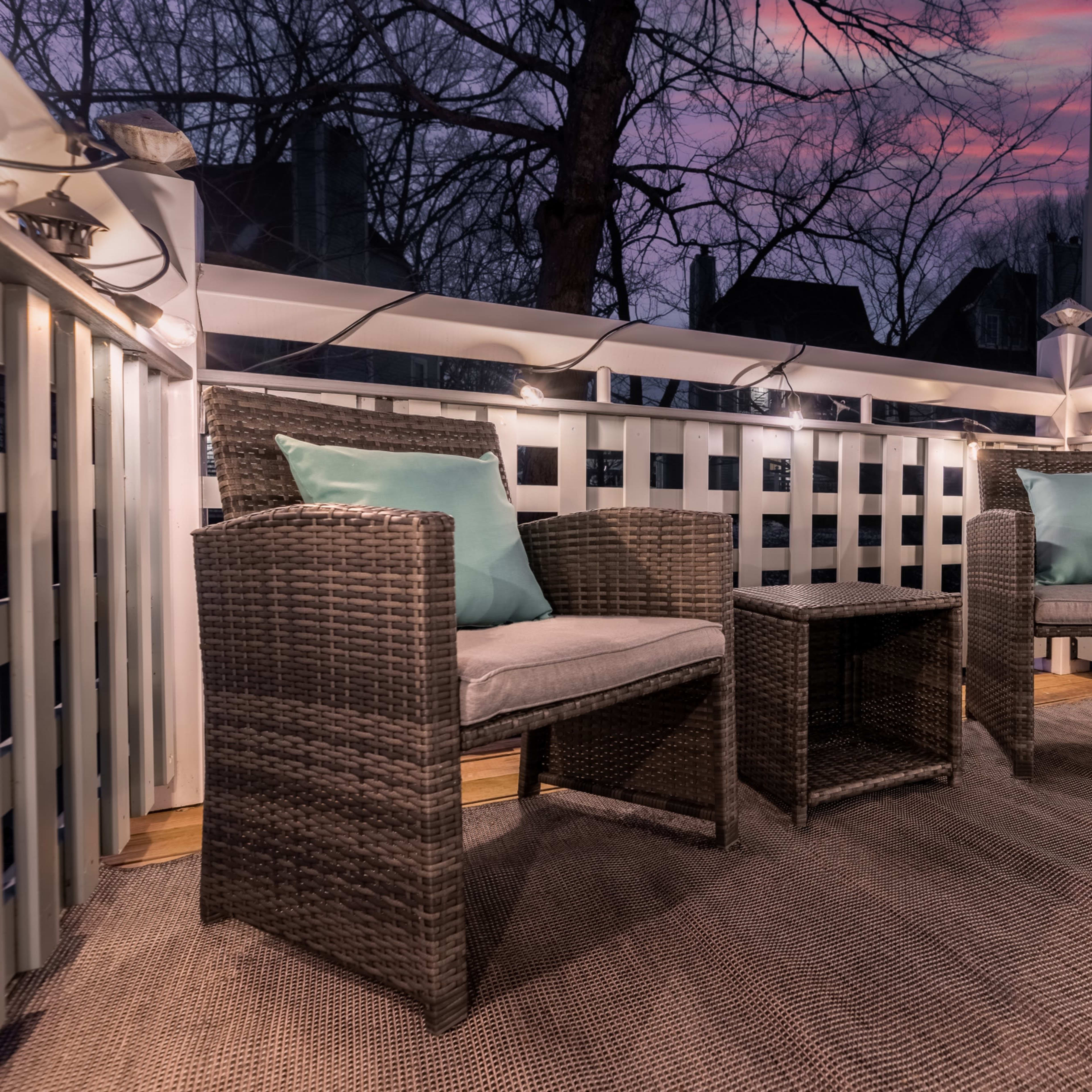 A small balcony features two wicker chairs and a table, surrounded by string lights, with a sunset sky visible in the background.