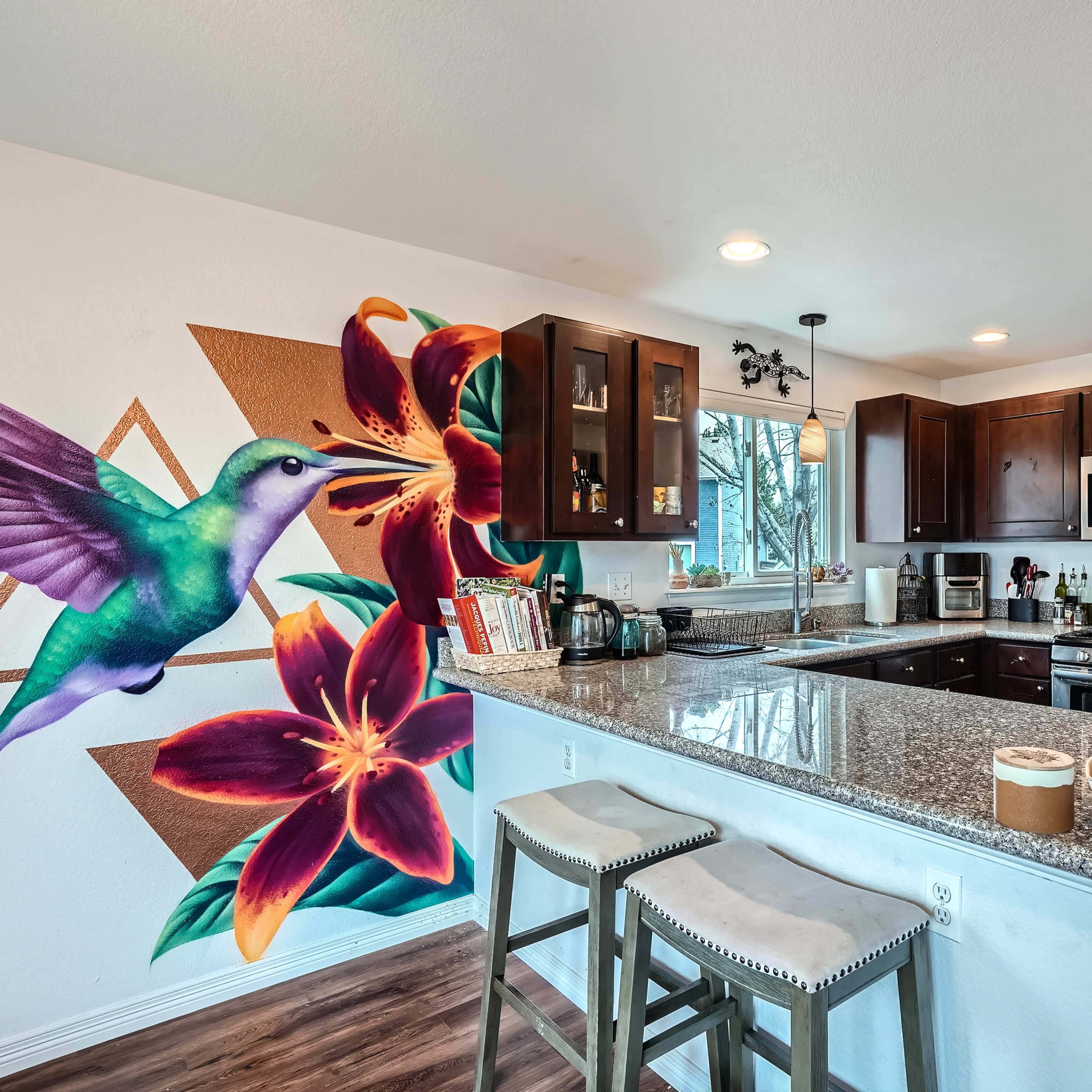 A modern kitchen features a large island with bar stools, dark wooden cabinets, and a colorful mural of a hummingbird and flowers on one wall.