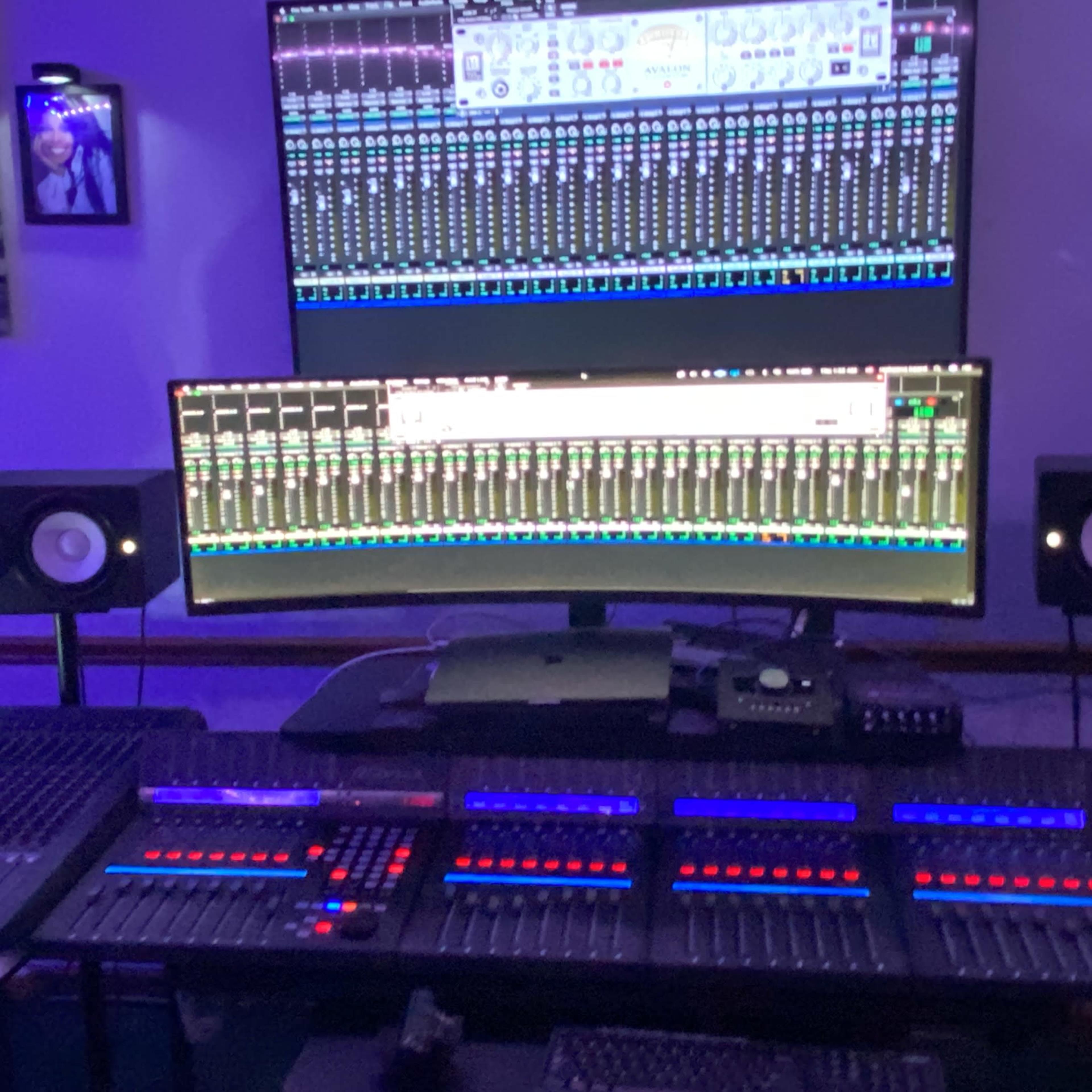 A music production studio with multiple large computer screens displaying audio mixing software, surrounded by speakers and a control panel with illuminated buttons.