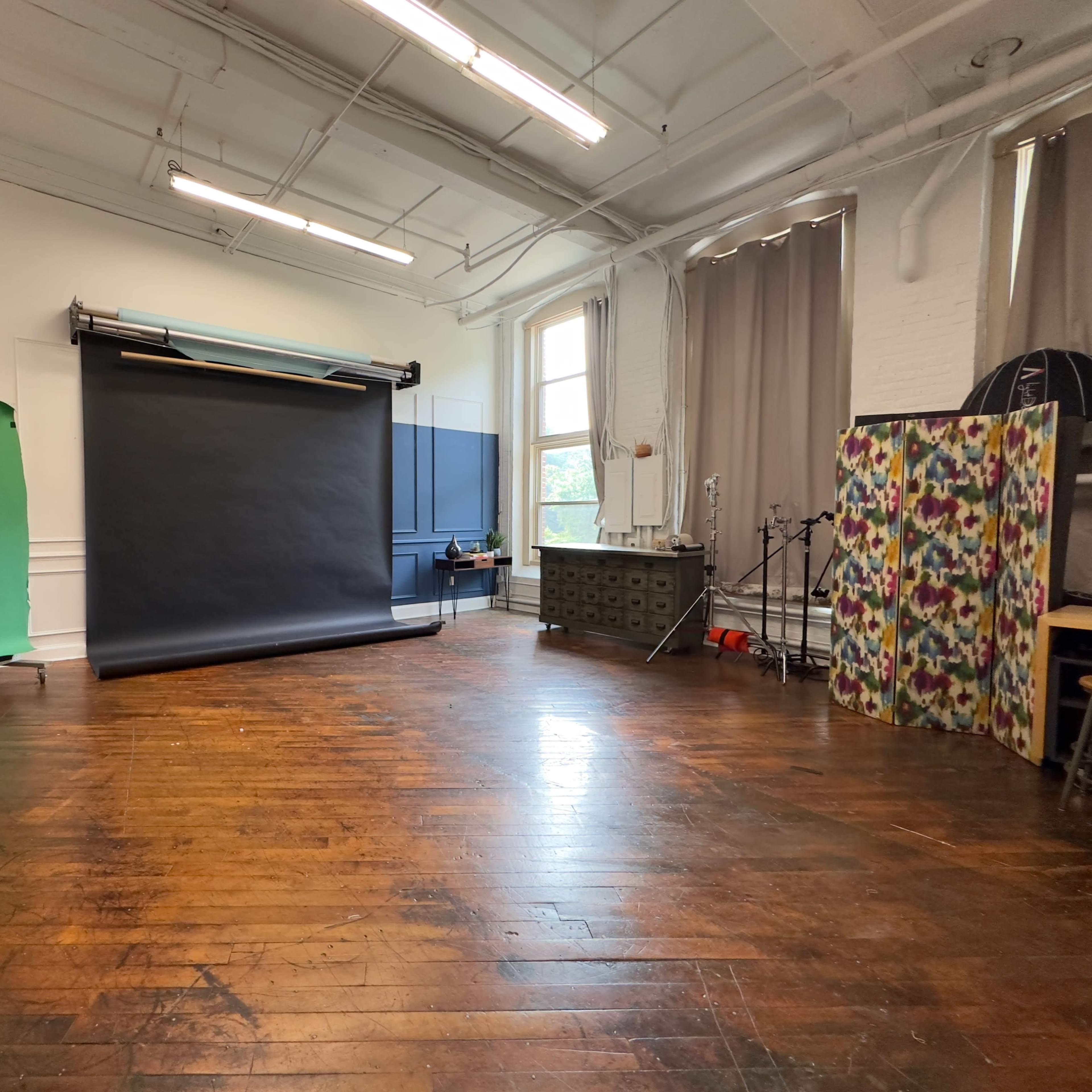 A photography studio with a wooden floor, a large black backdrop, a green screen, and various equipment arranged around the space.