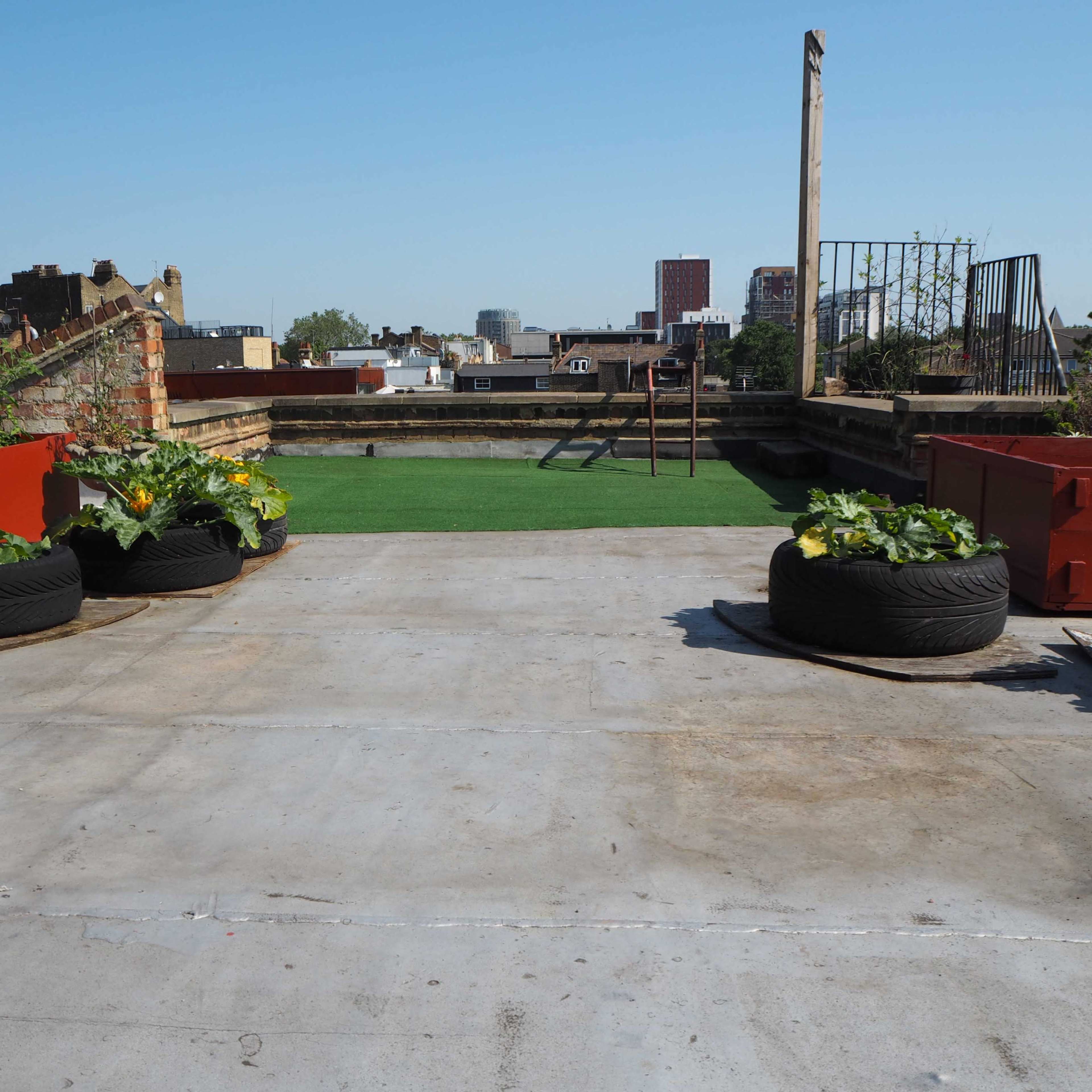 A rooftop garden features green plants in tire planters, with a grassy area and city buildings visible in the background.