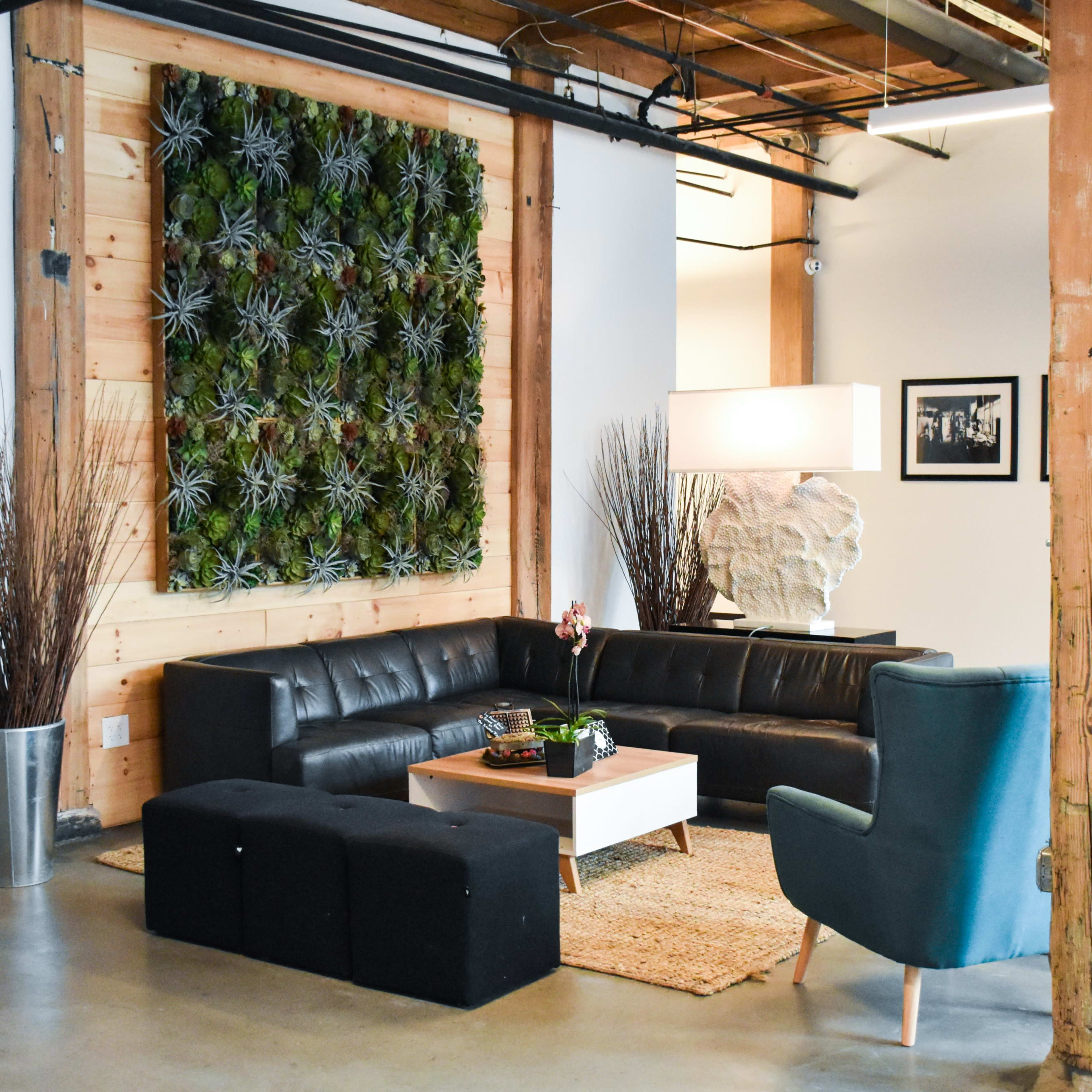 The scene features a modern lounge area with a black leather sofa, teal armchairs, a wooden coffee table, and a vertical wall of greenery, all set against a backdrop of exposed wooden beams.