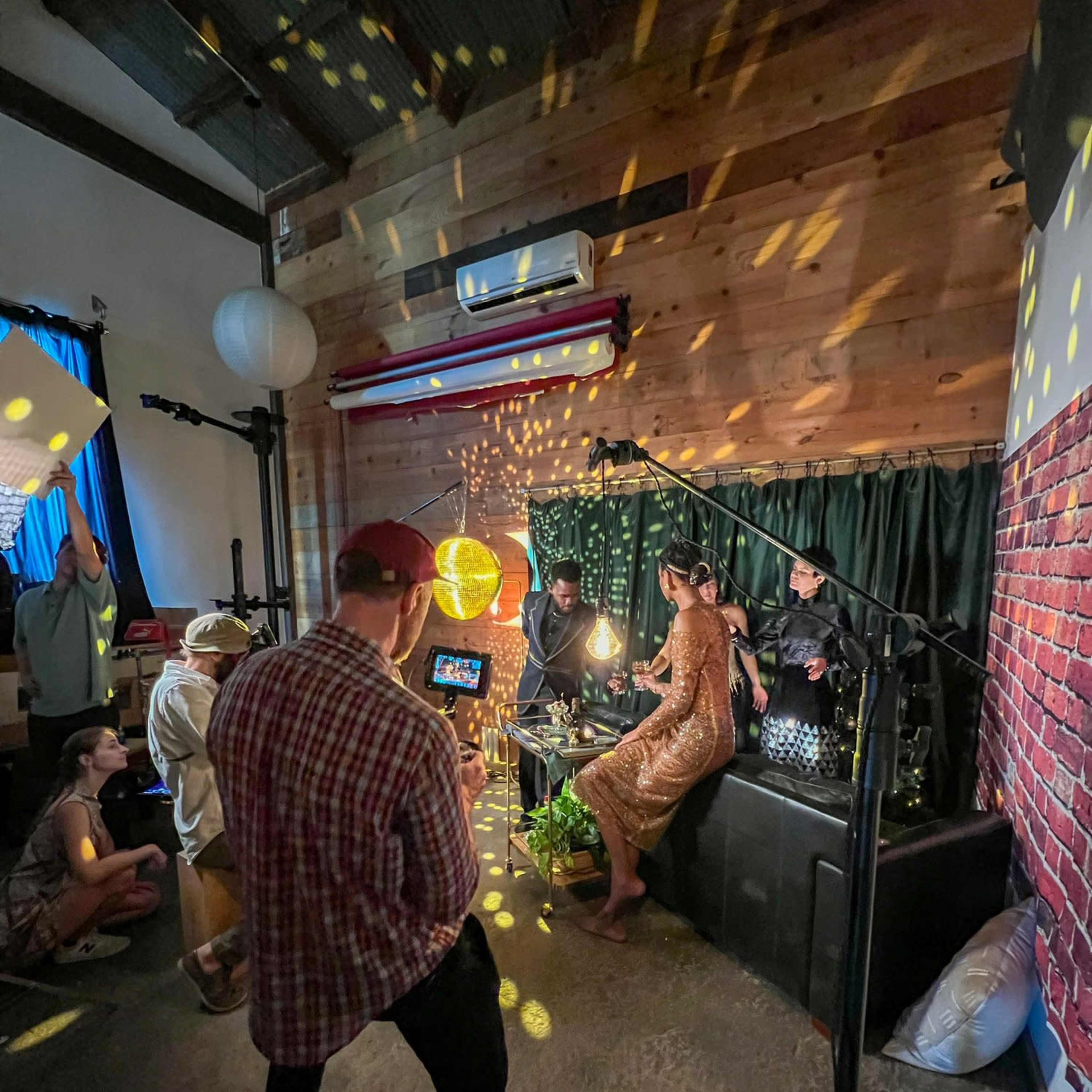 A group of people is gathered in a dimly lit room with wooden walls, engaged in a photoshoot or video production involving a model in a shiny dress and decorative lighting effects.