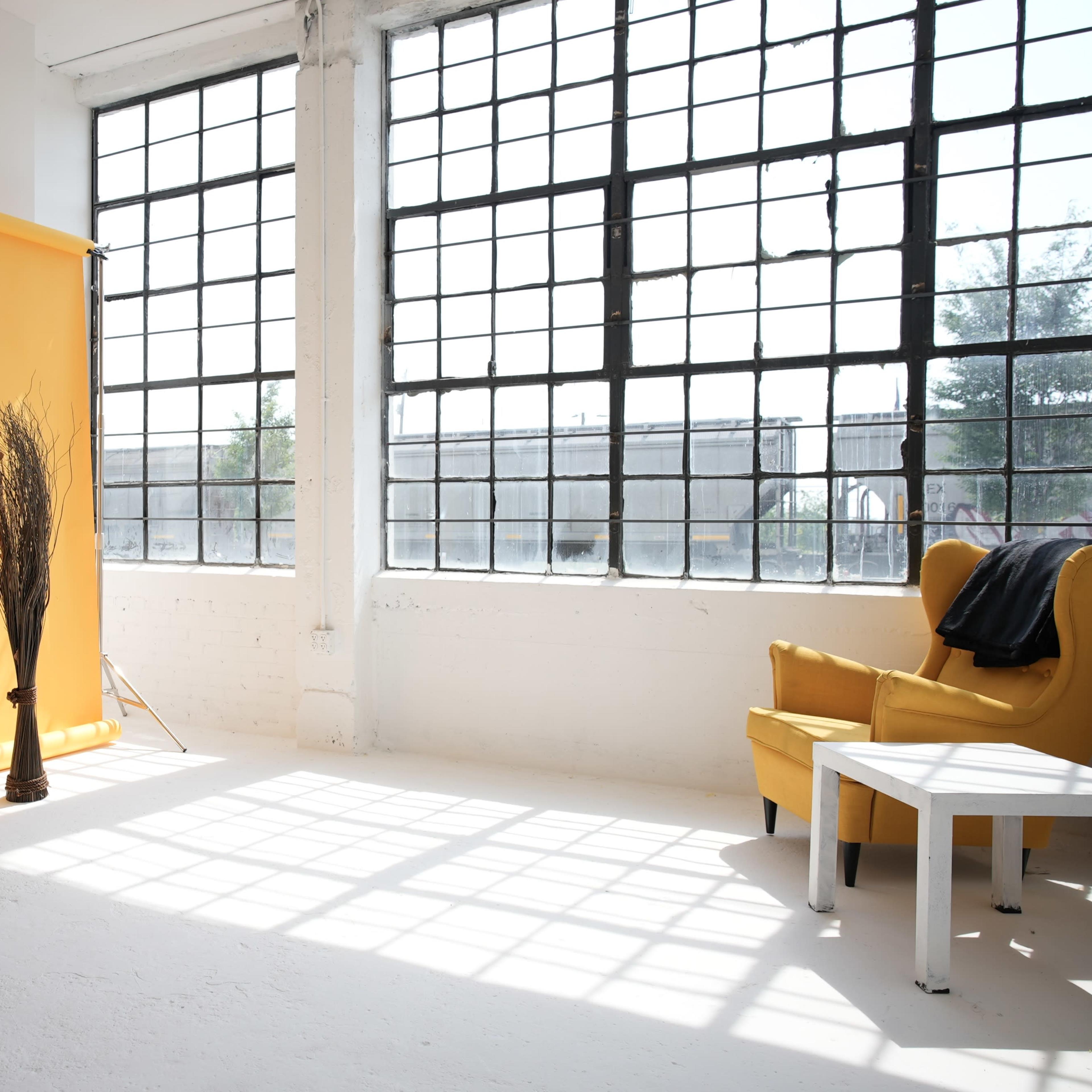 A bright studio features a yellow backdrop, a wooden stool, a tall plant, and a yellow armchair beside a white table, with large windows casting shadows on the floor.