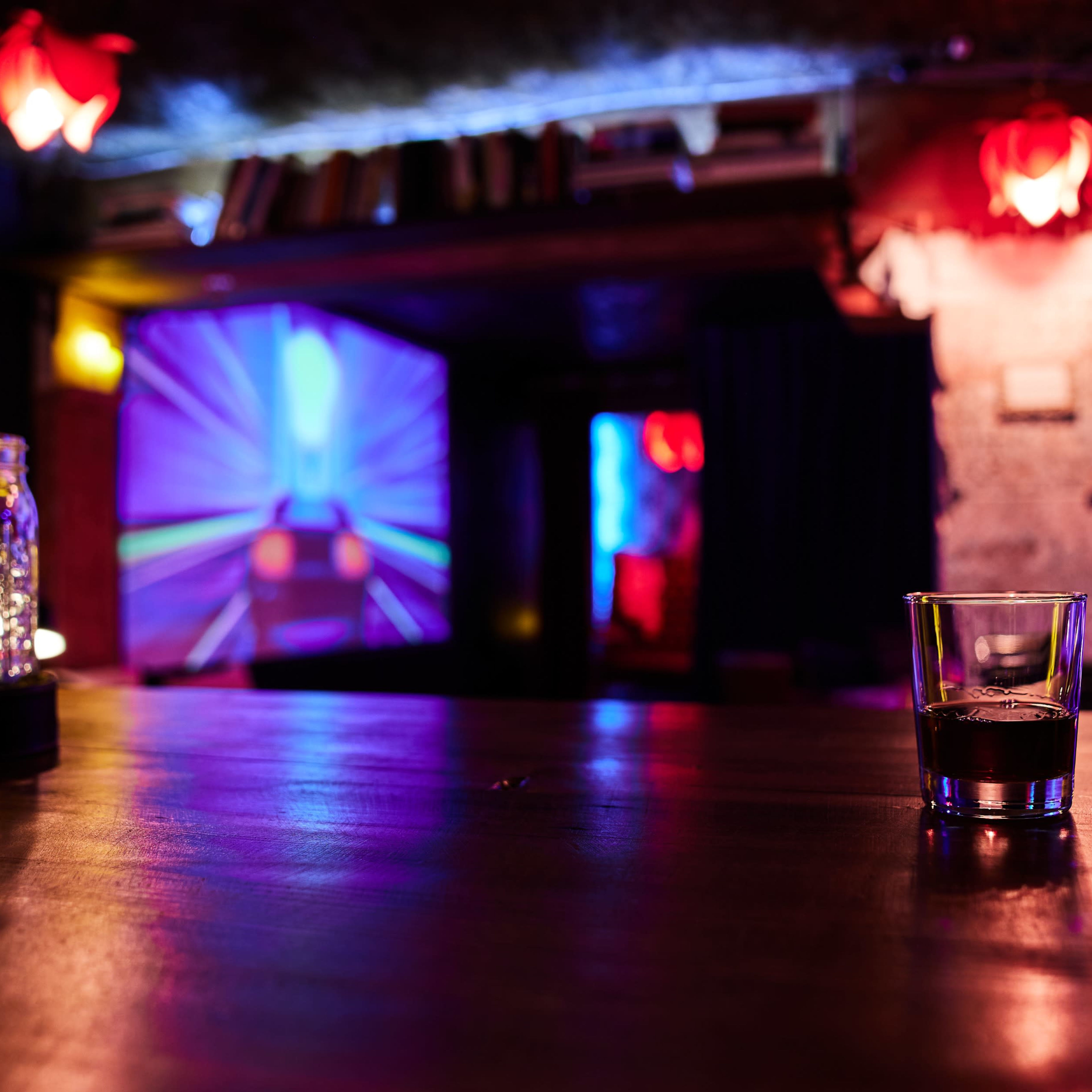 A glass sits on a wooden bar in a dimly lit room with colorful lights illuminating the walls and a projection of a vibrant scene.