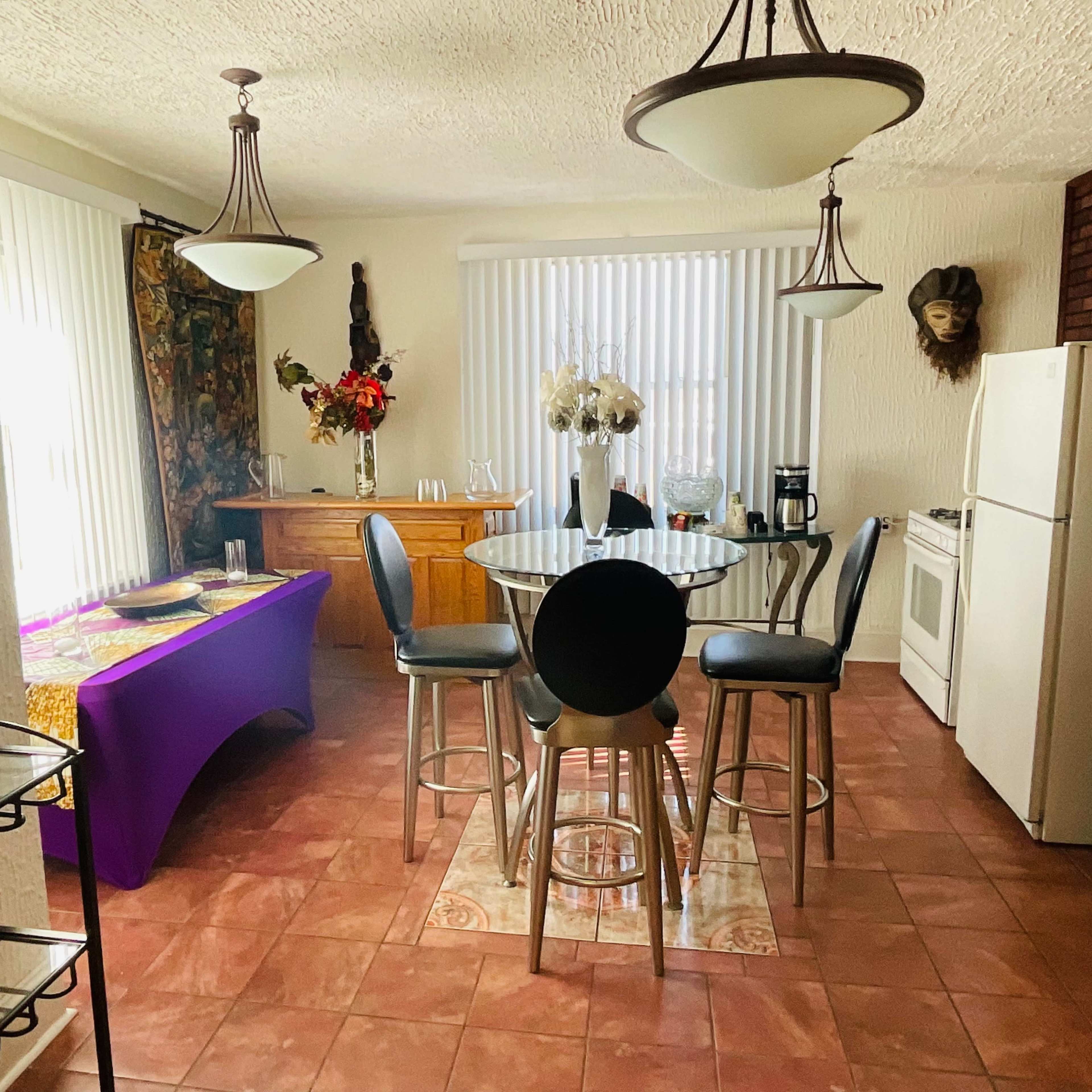 A kitchen dining area features a round glass table with high-backed chairs, a sideboard, and a white refrigerator, along with large windows covered by vertical blinds.