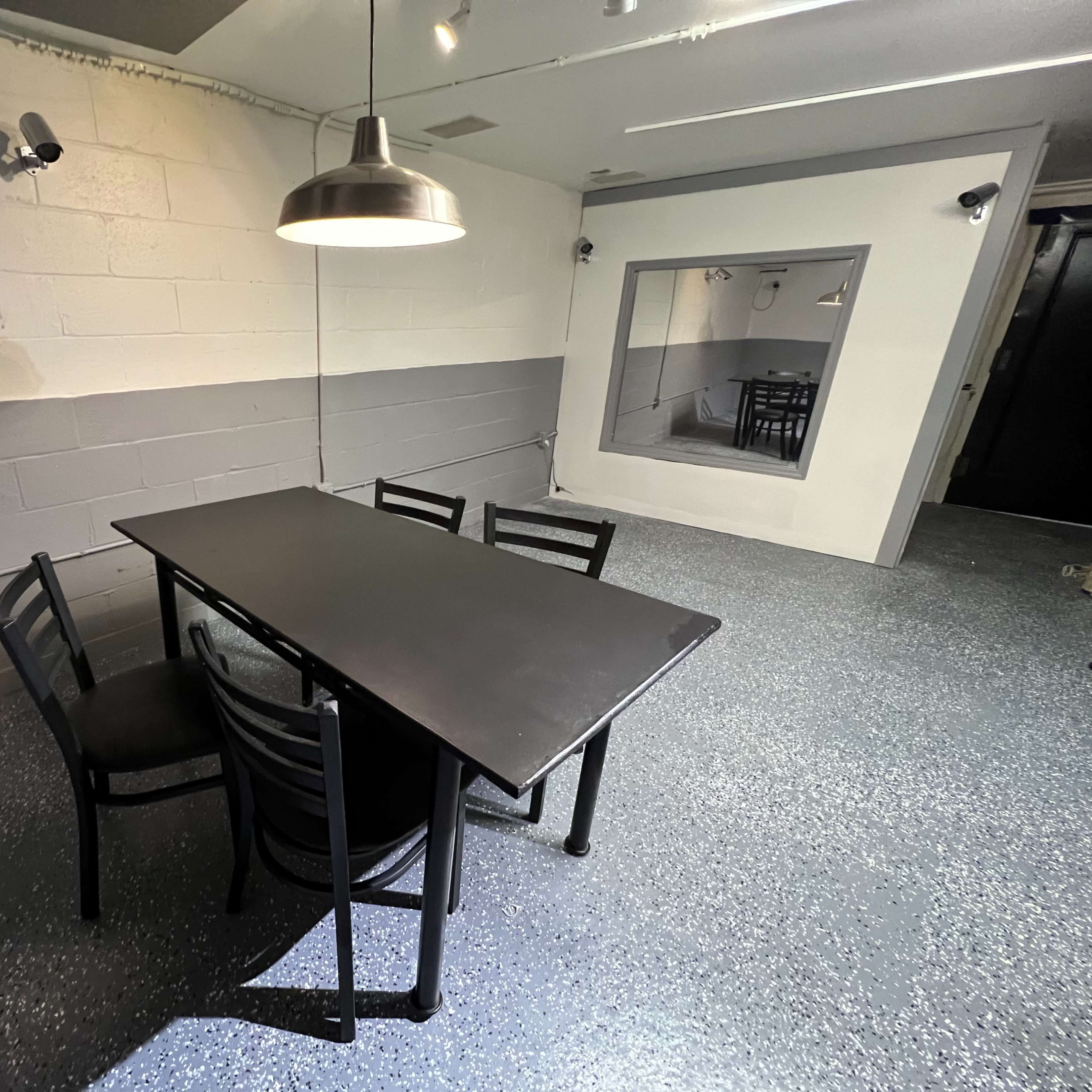 A black table surrounded by four chairs is positioned in a gray-walled room with a large mirror on one side.