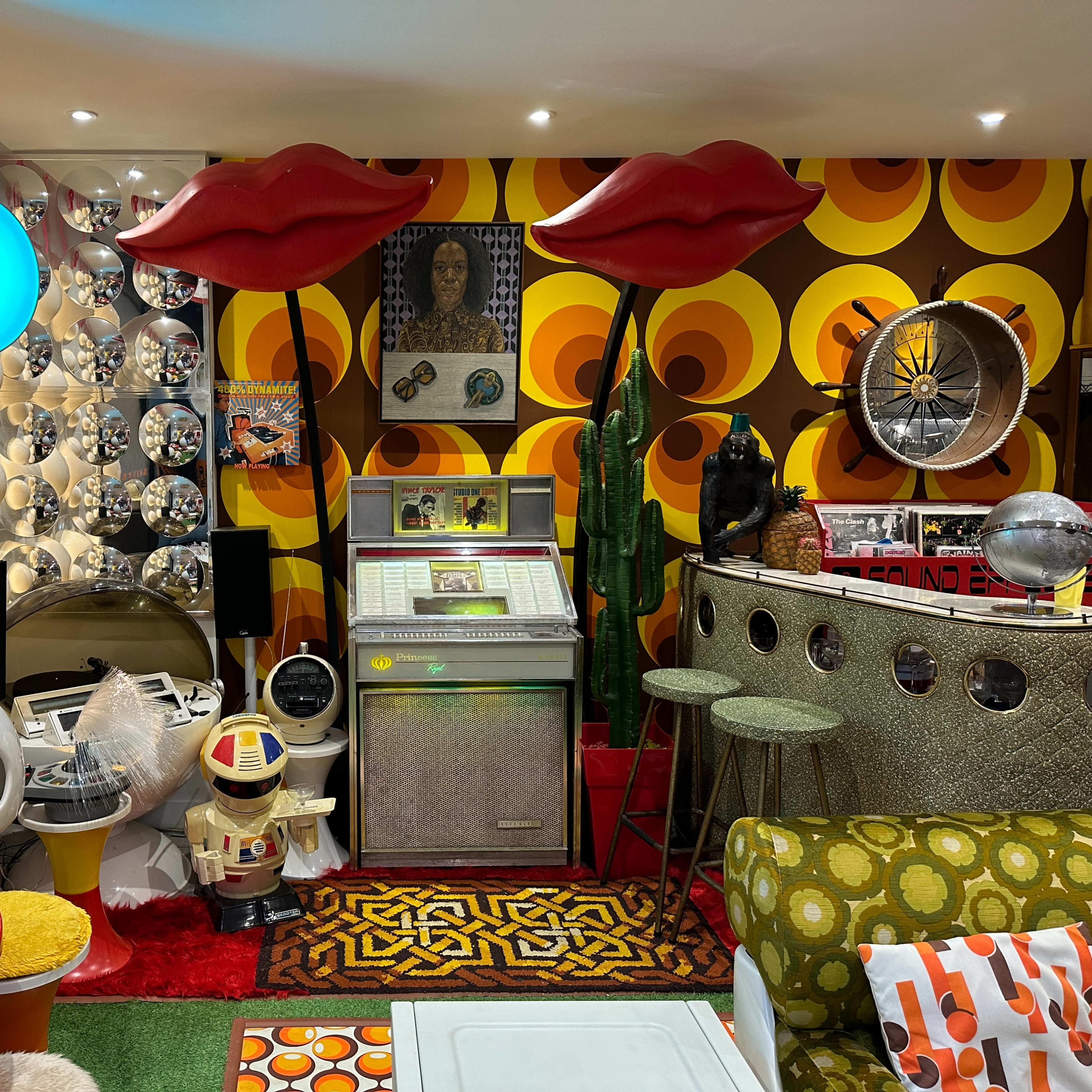 A vibrant retro-themed room filled with colorful furniture, vintage decor, and unique art pieces, including large lip-shaped structures and a vintage jukebox.