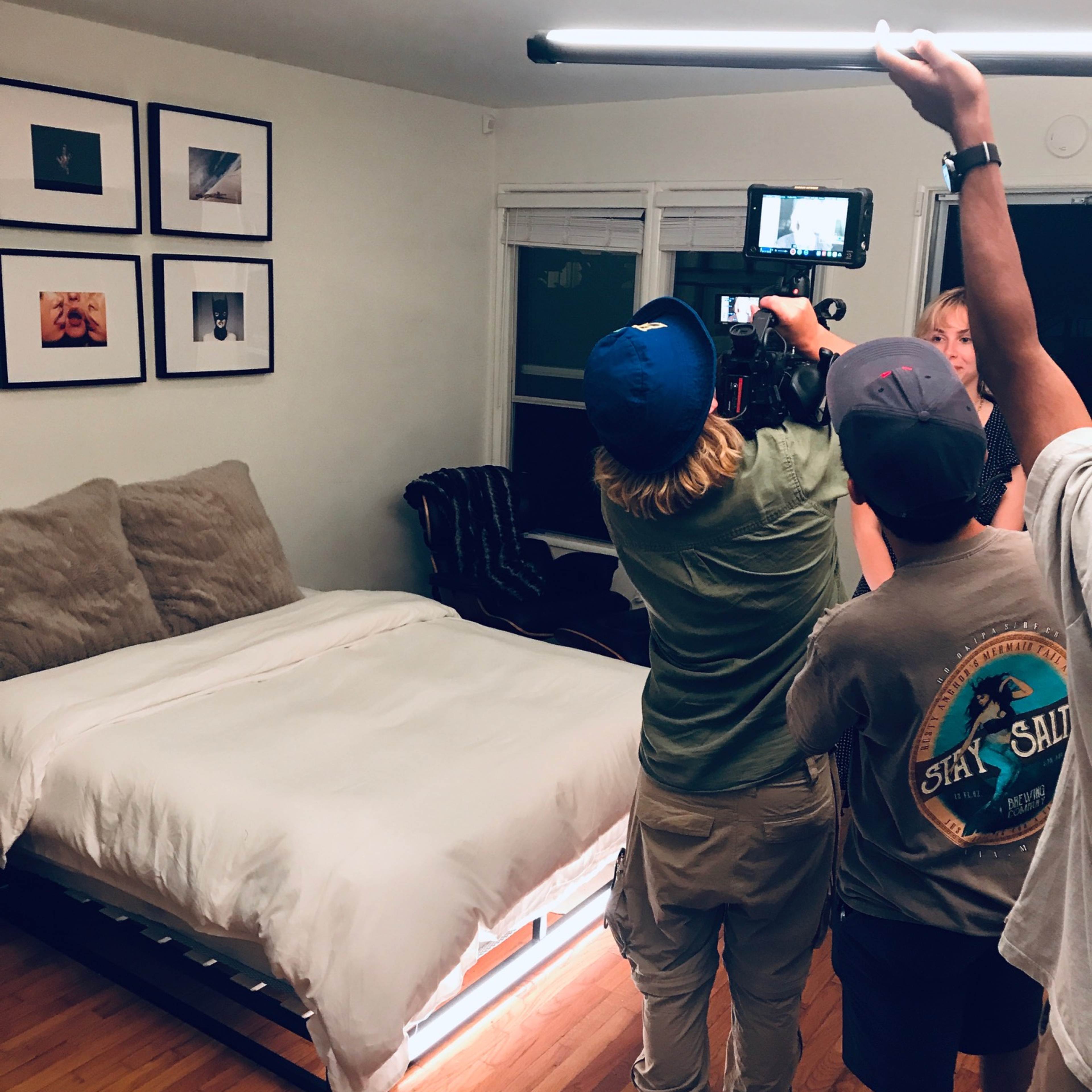 A crew is filming in a well-lit bedroom with a large bed, while one person holds a camera on a tripod and two others assist with lighting.