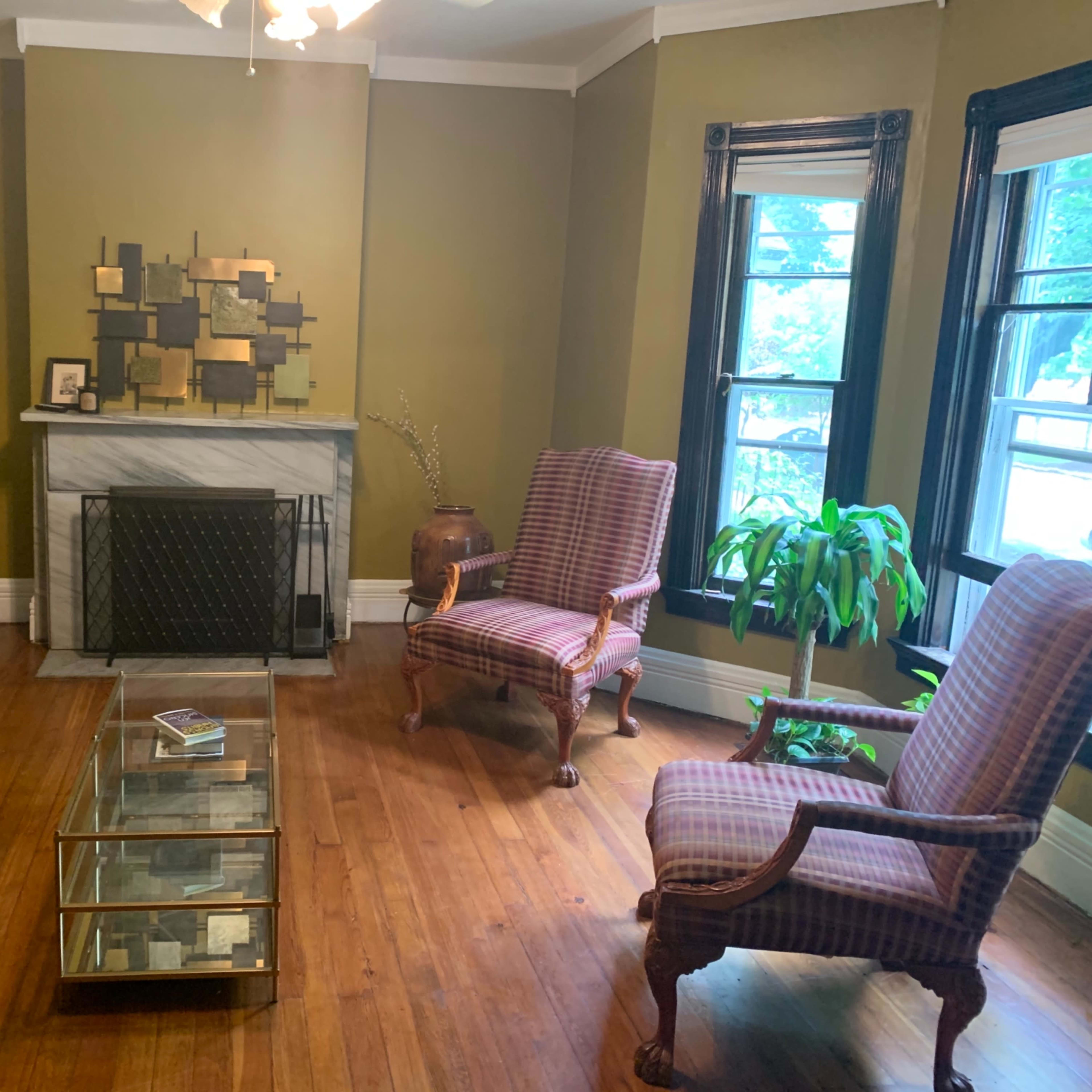A living room with a gray sofa, two patterned armchairs, a glass coffee table, and a marble fireplace against a green wall.