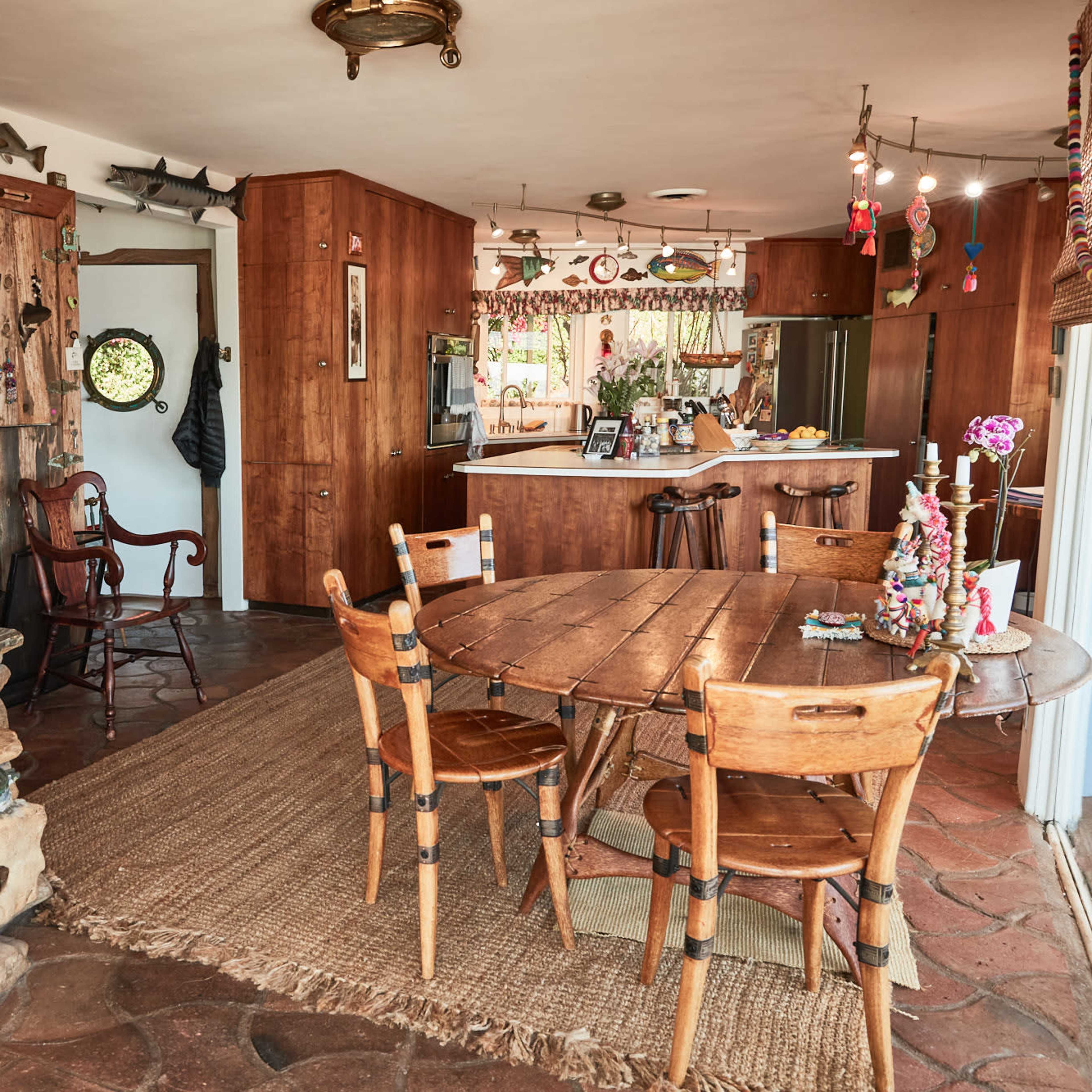 A rustic dining area with a round wooden table, surrounding chairs, and a colorful decor featuring wall hangings and various ornaments.