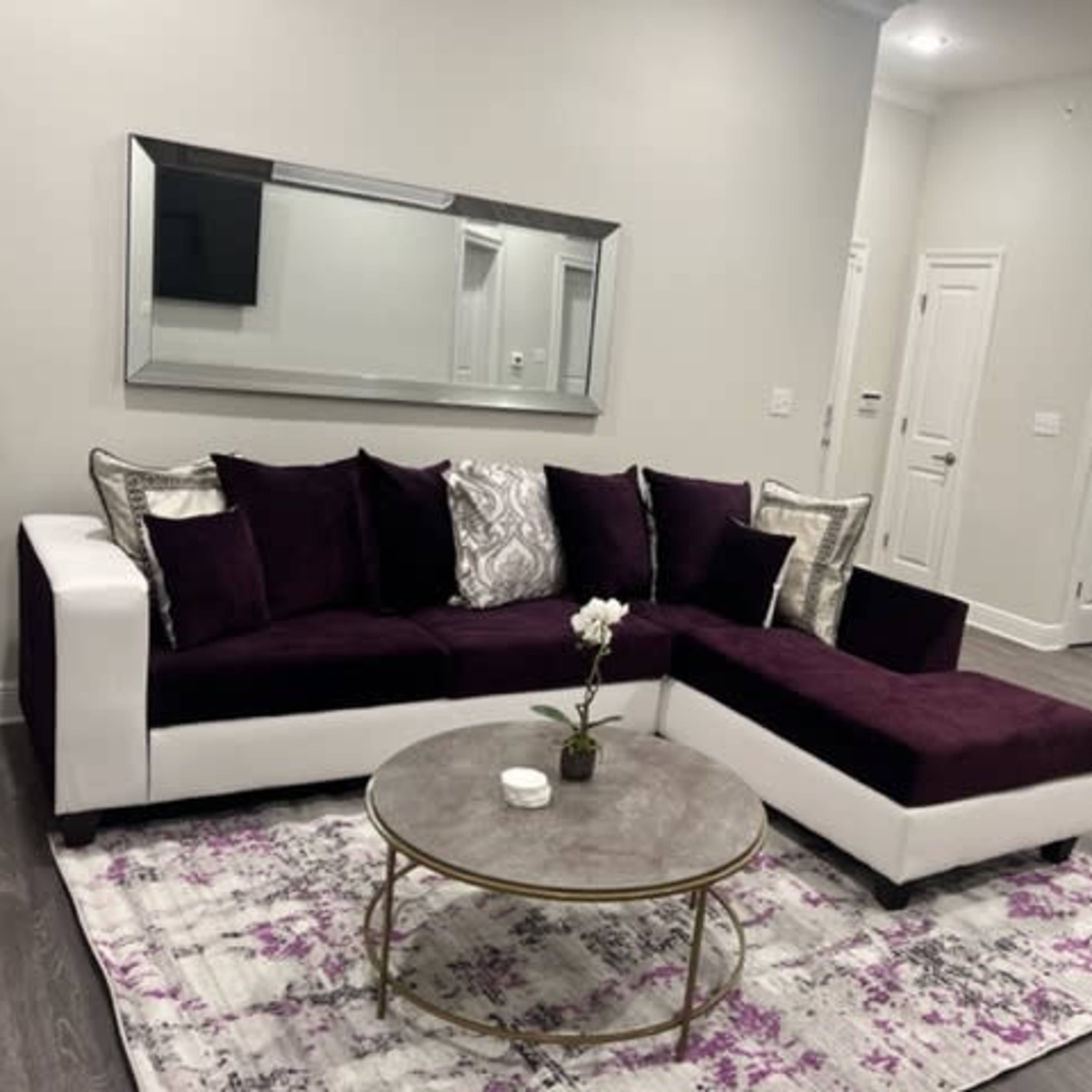 A modern living room featuring a purple and white sectional sofa, a round coffee table, and a mirror on the wall, with a kitchen visible in the background.