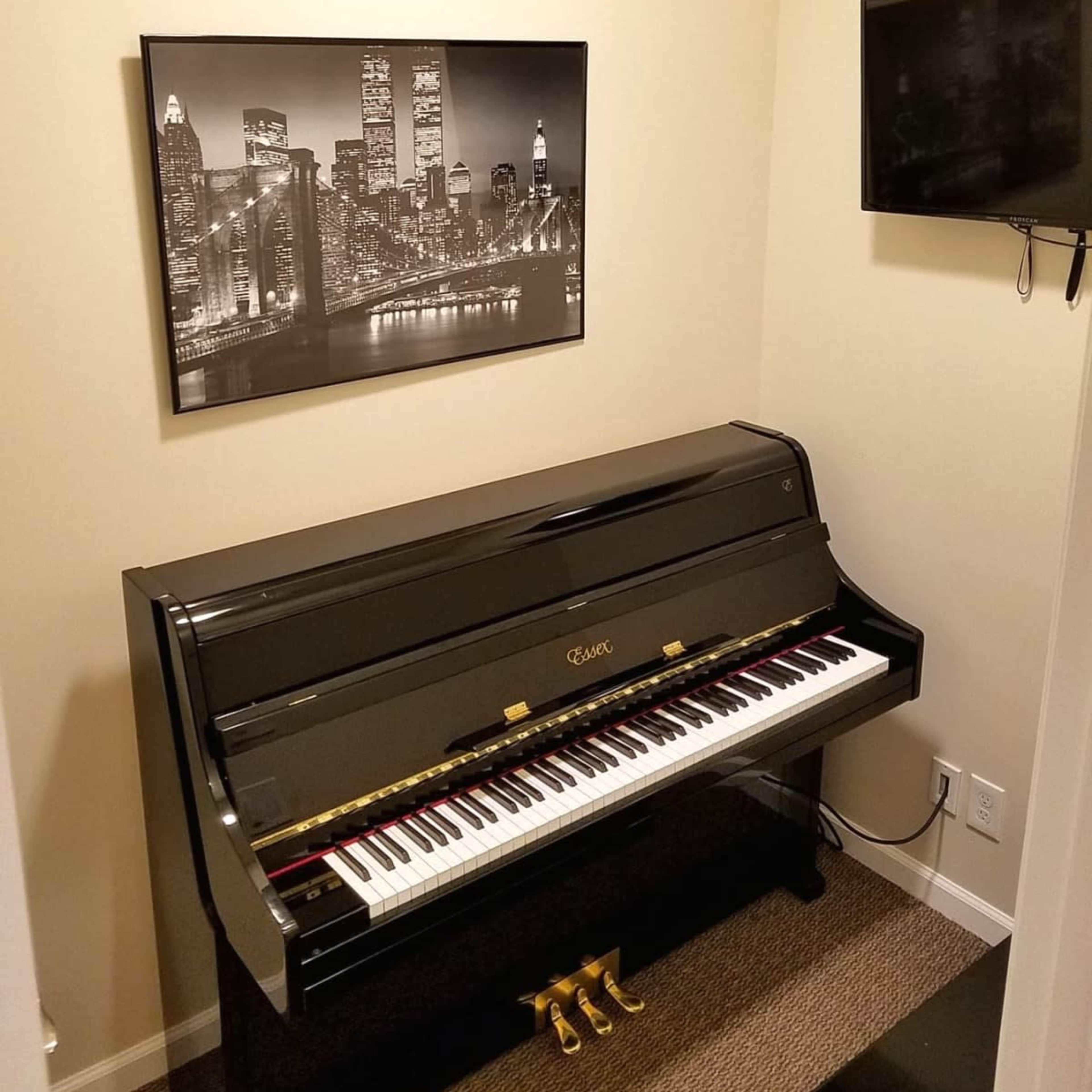 A black piano is positioned against a wall in a small room, featuring a black-and-white cityscape photograph and a mounted television nearby.