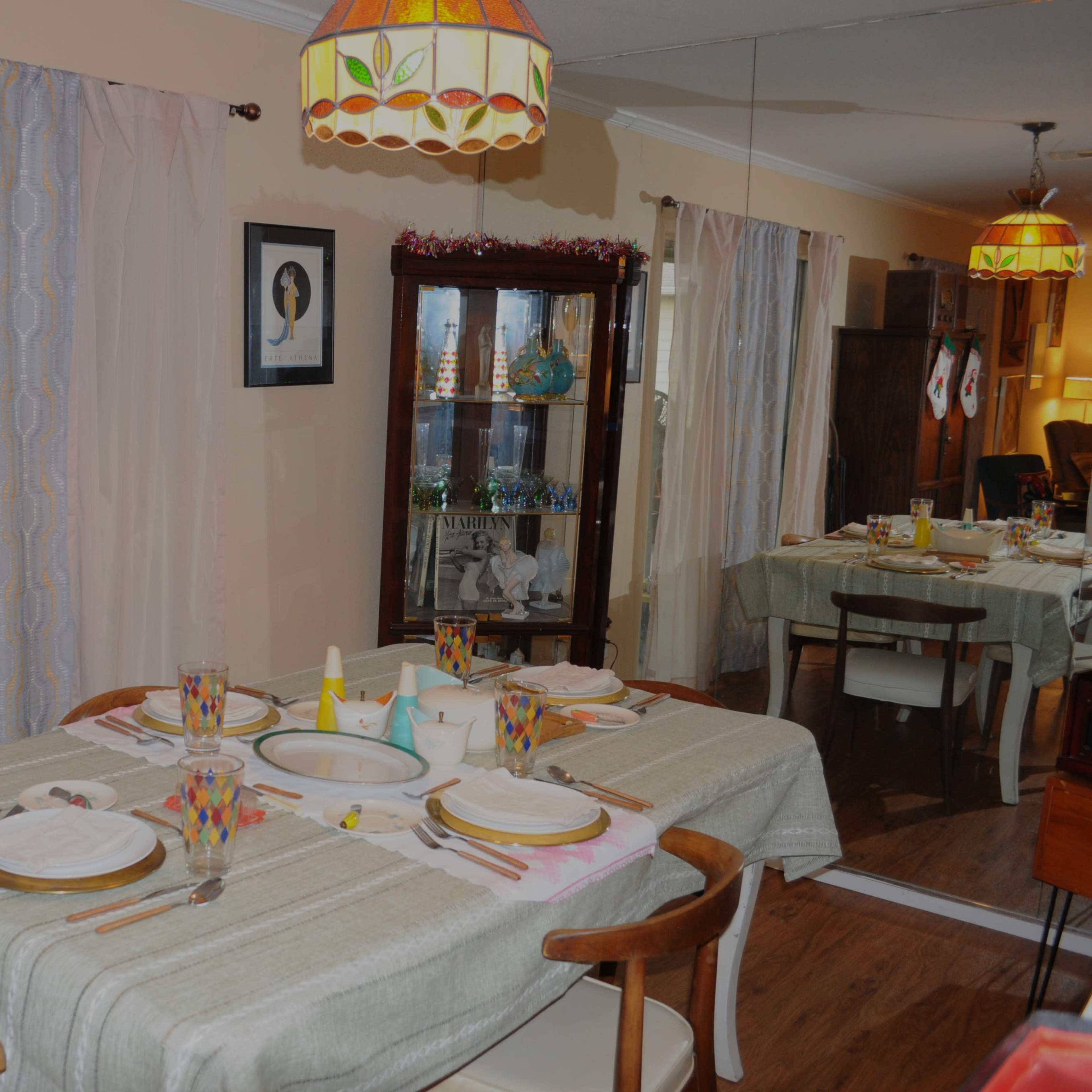 A dining room with a table set for a meal, featuring plates, glasses, and a centrally placed bowl, surrounded by chairs and a cabinet displaying decorative items.