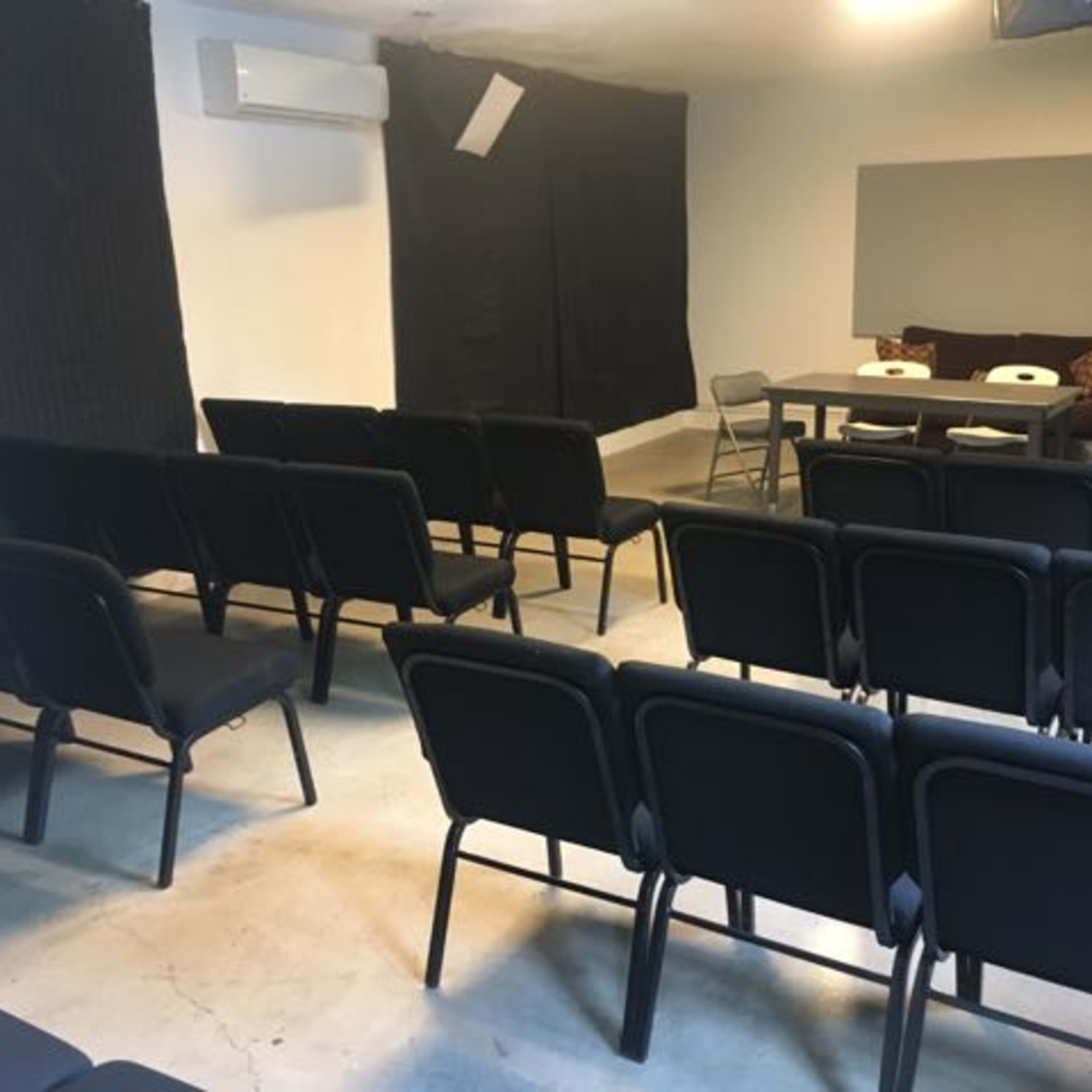 A room with a row of black chairs facing a table and a wall, with additional chairs arranged to the side.