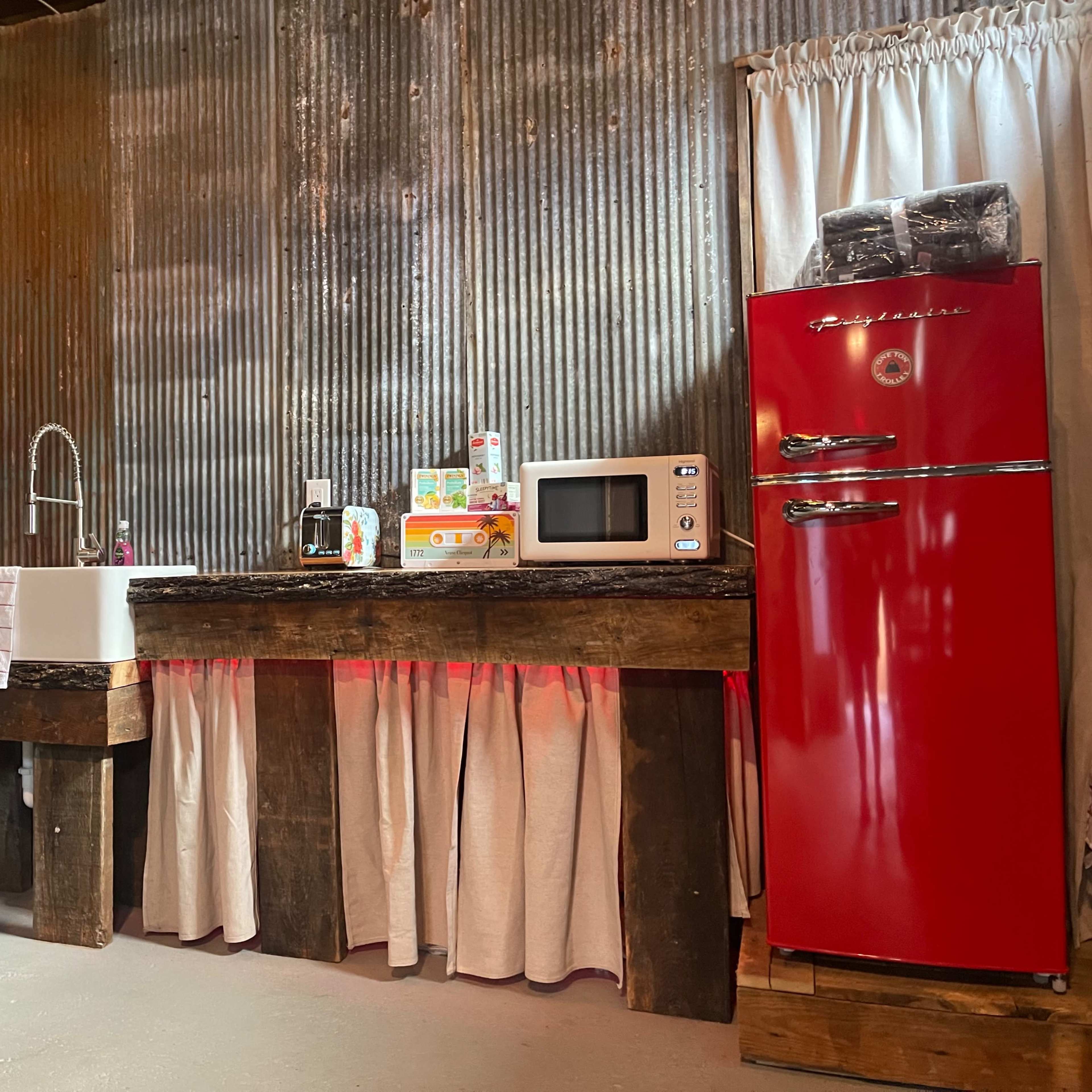 A rustic kitchen featuring a large farmhouse sink, a red retro-style refrigerator, a microwave, and wooden countertops with a curtain below.