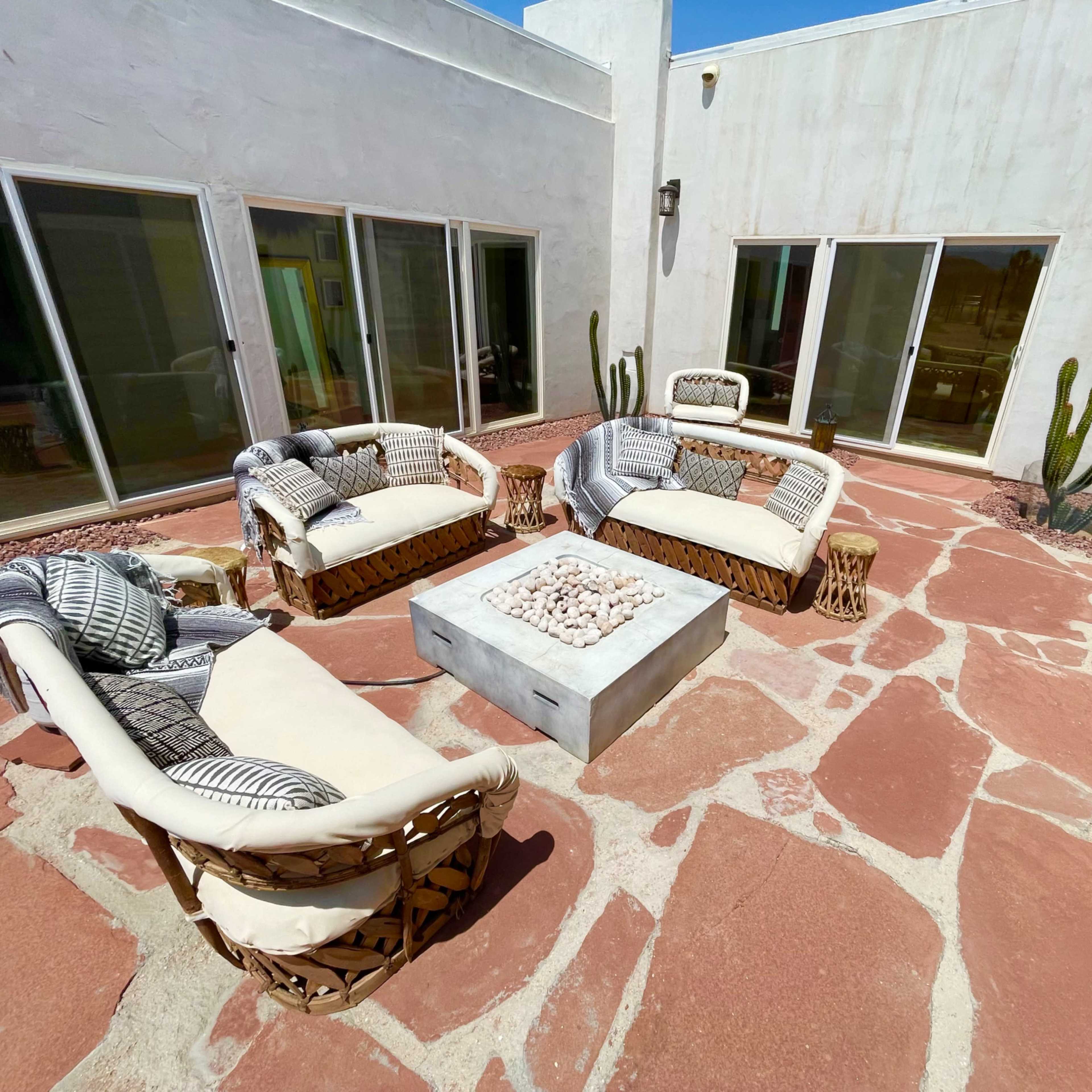 A patio area features four wicker chairs arranged around a rectangular stone fire pit, with large windows and cacti in the background.