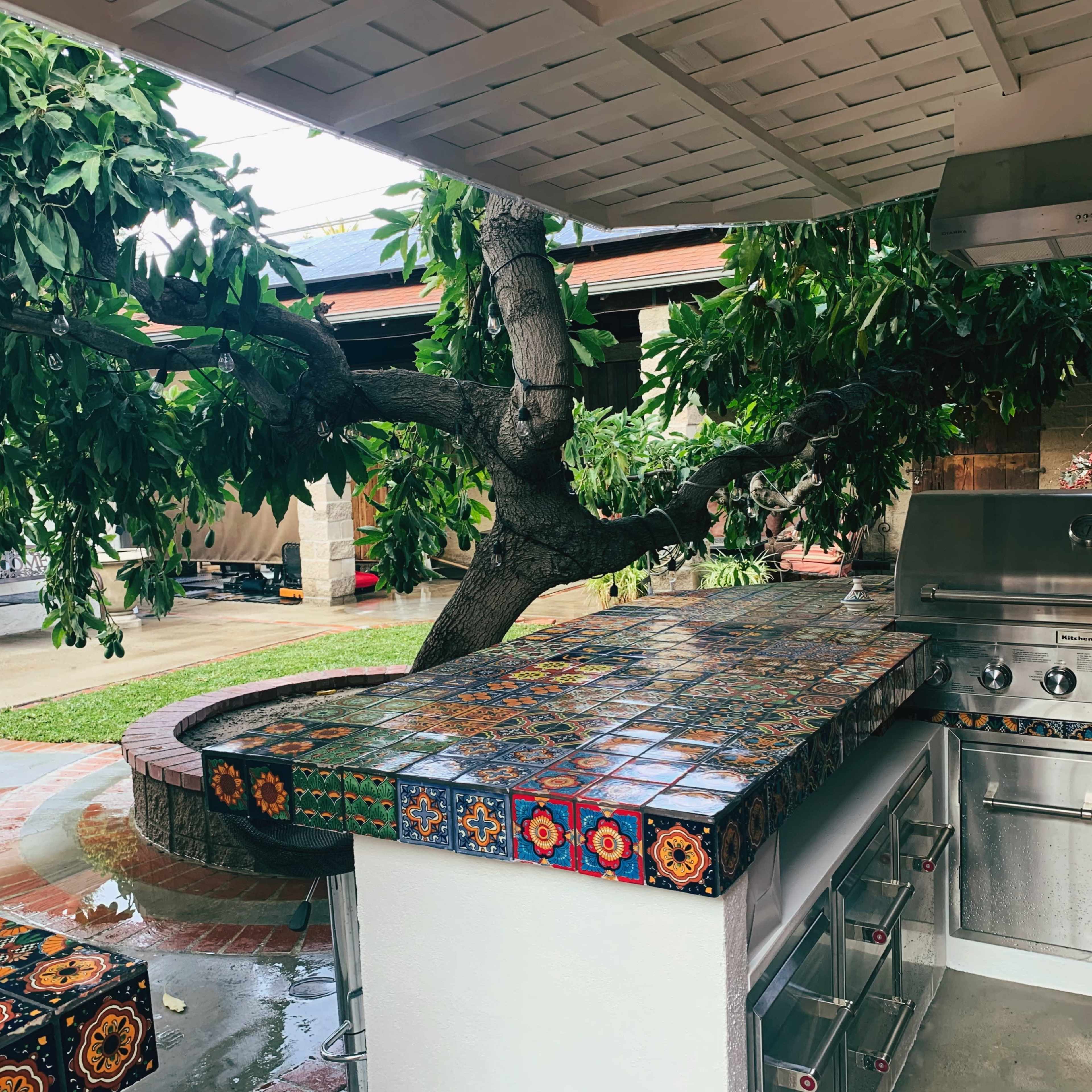 An outdoor kitchen features a tiled bar area and a grill, surrounded by a tree and green foliage.