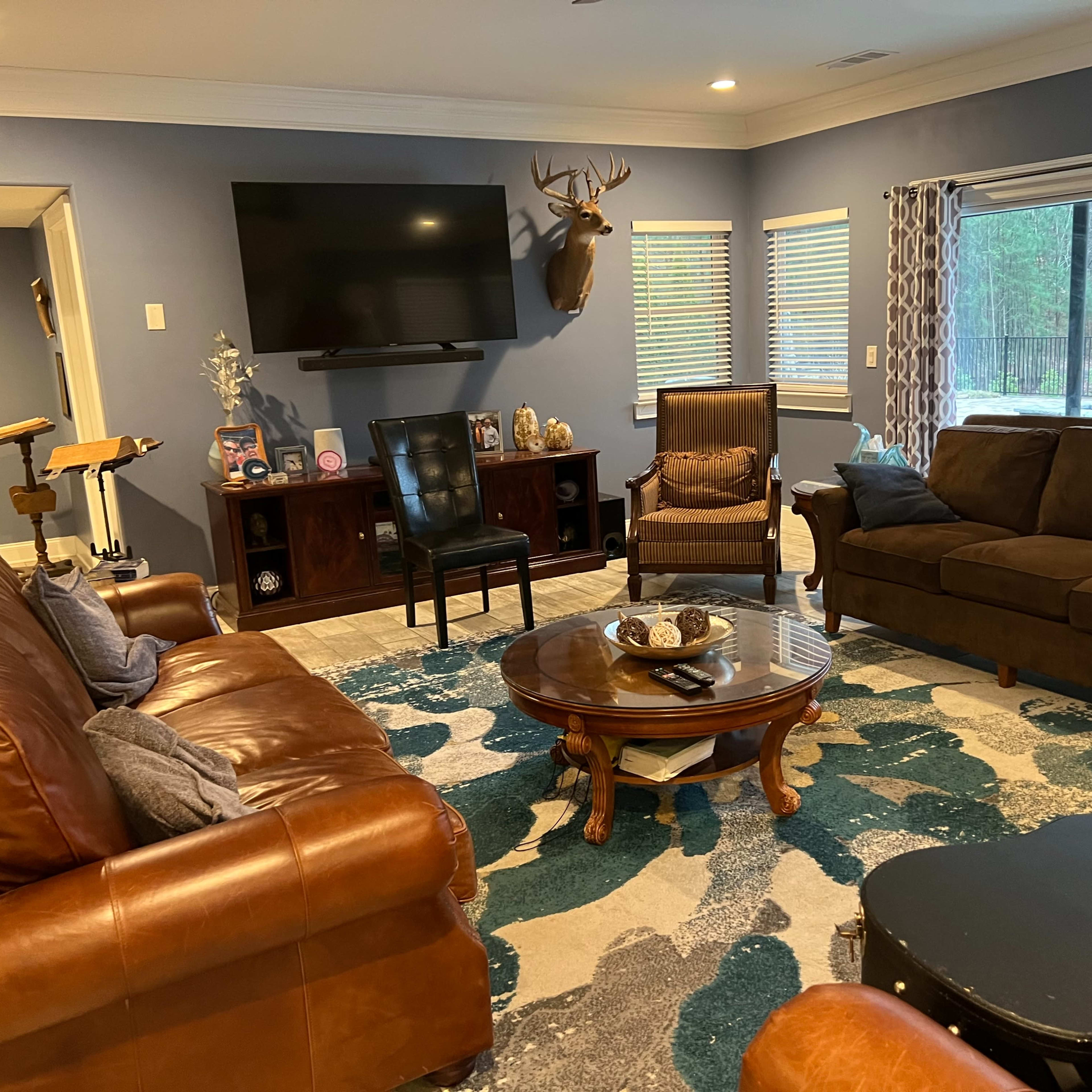 A living room with a large television mounted on the wall, a mounted deer head, and a mix of leather and fabric furniture arranged around a coffee table on a patterned rug.
