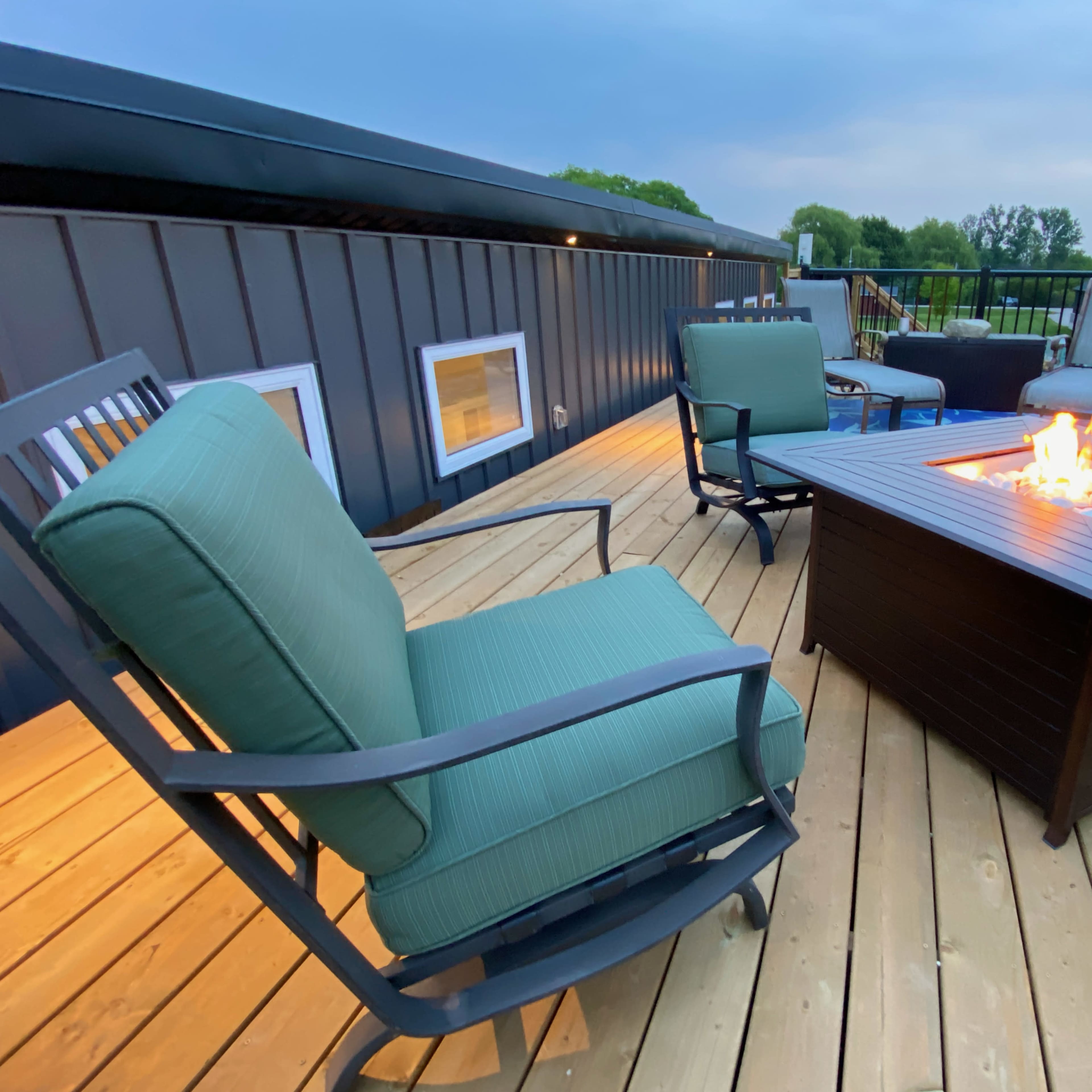 A rectangular fire pit with a flame sits on a wooden deck surrounded by green chairs.