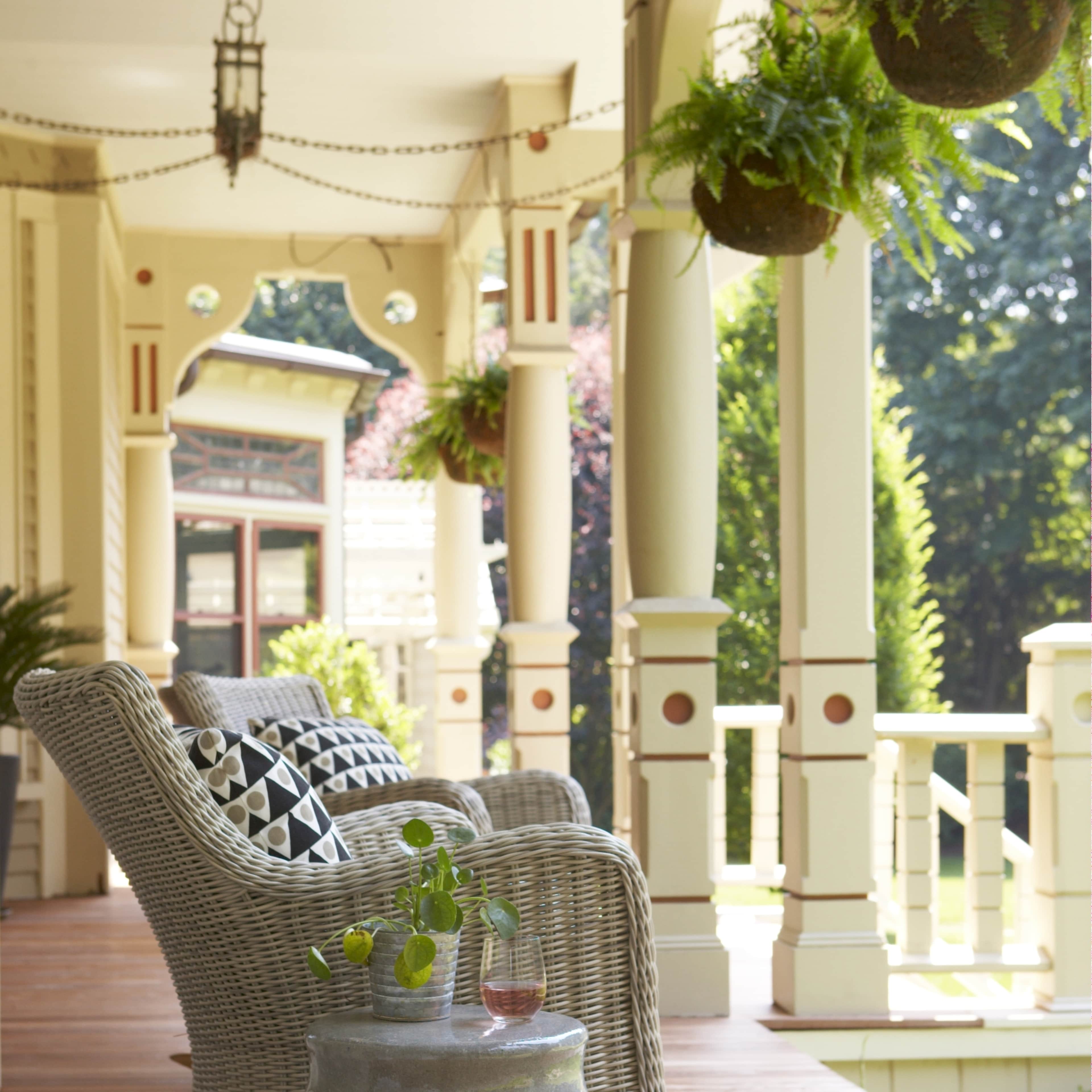 A wicker rocking chair sits on a wooden porch beside a round, textured side table with a small plant and a drink.