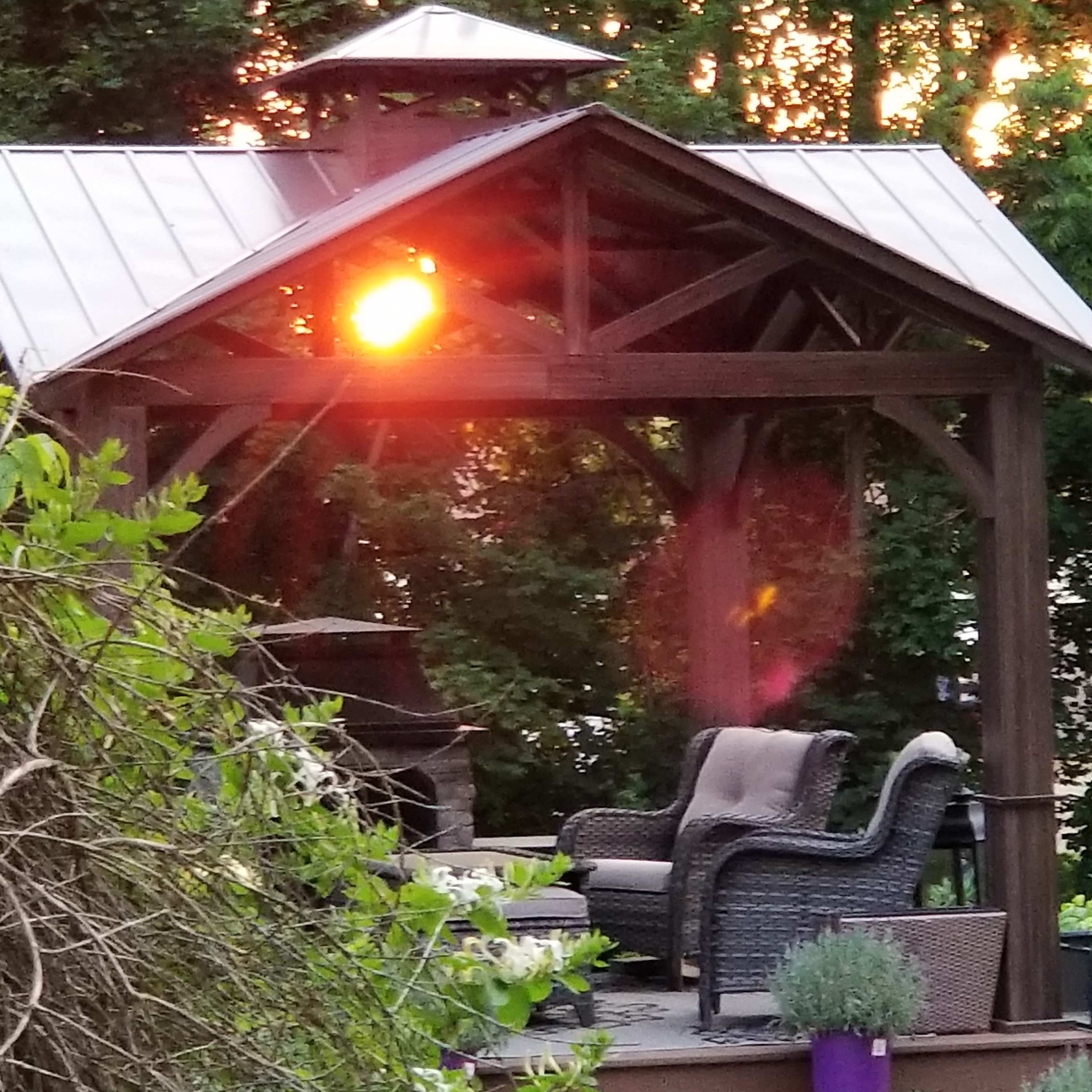 A wooden gazebo with a metal roof features a wicker chair and is illuminated by the setting sun in the background.
