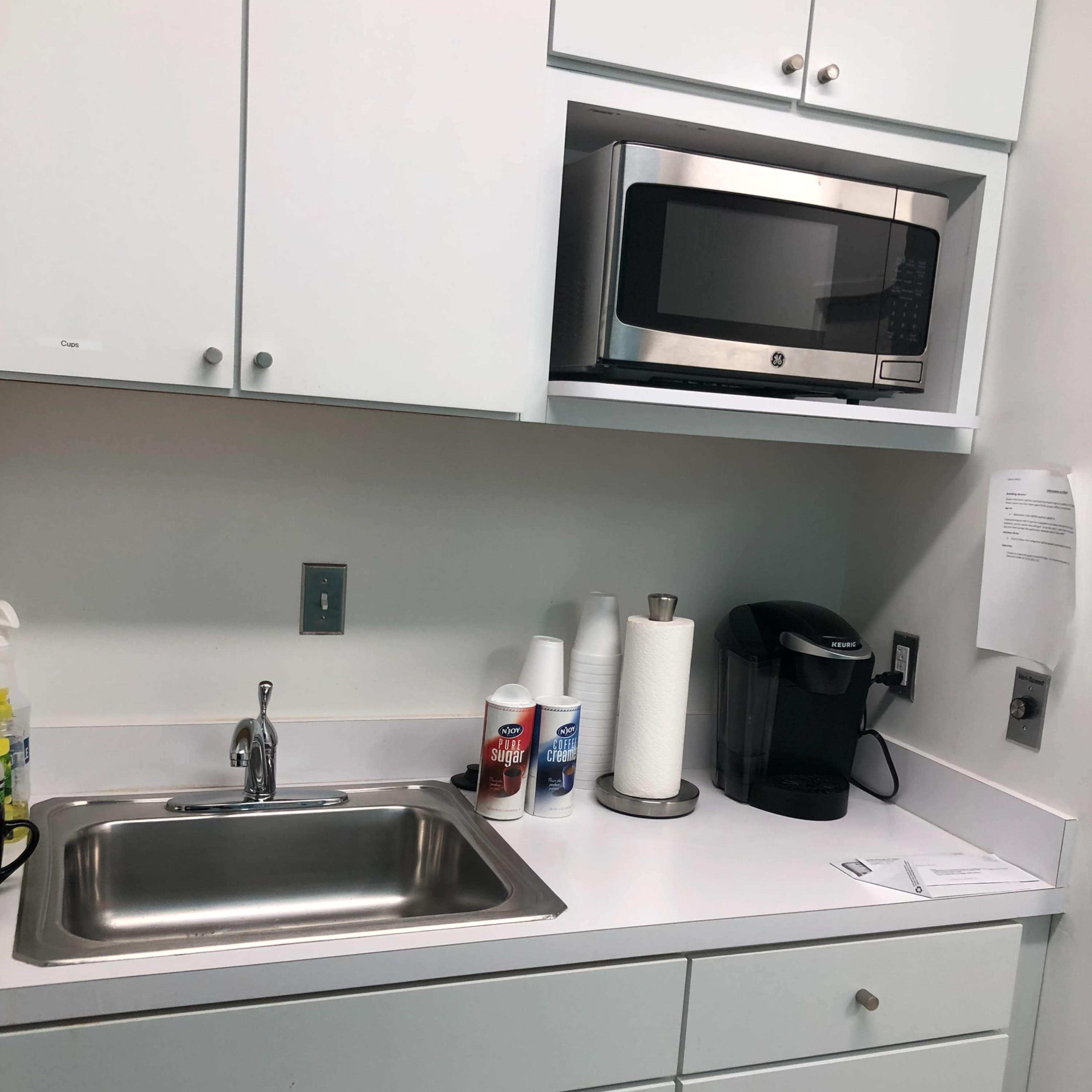 A clean kitchenette featuring a sink, a microwave mounted above, and a coffee maker on the countertop with cleaning supplies nearby.