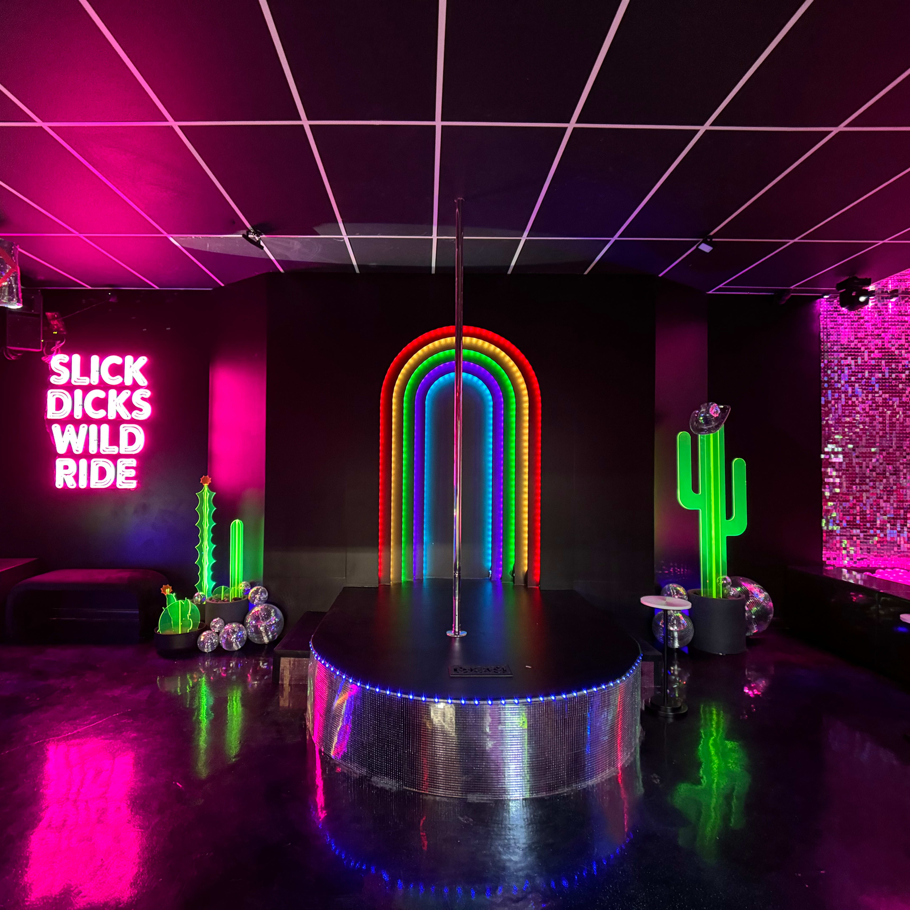A vibrant nightclub interior with neon lights, including a rainbow arch, a pole in the center, and decorative elements like cactus figures on the walls.
