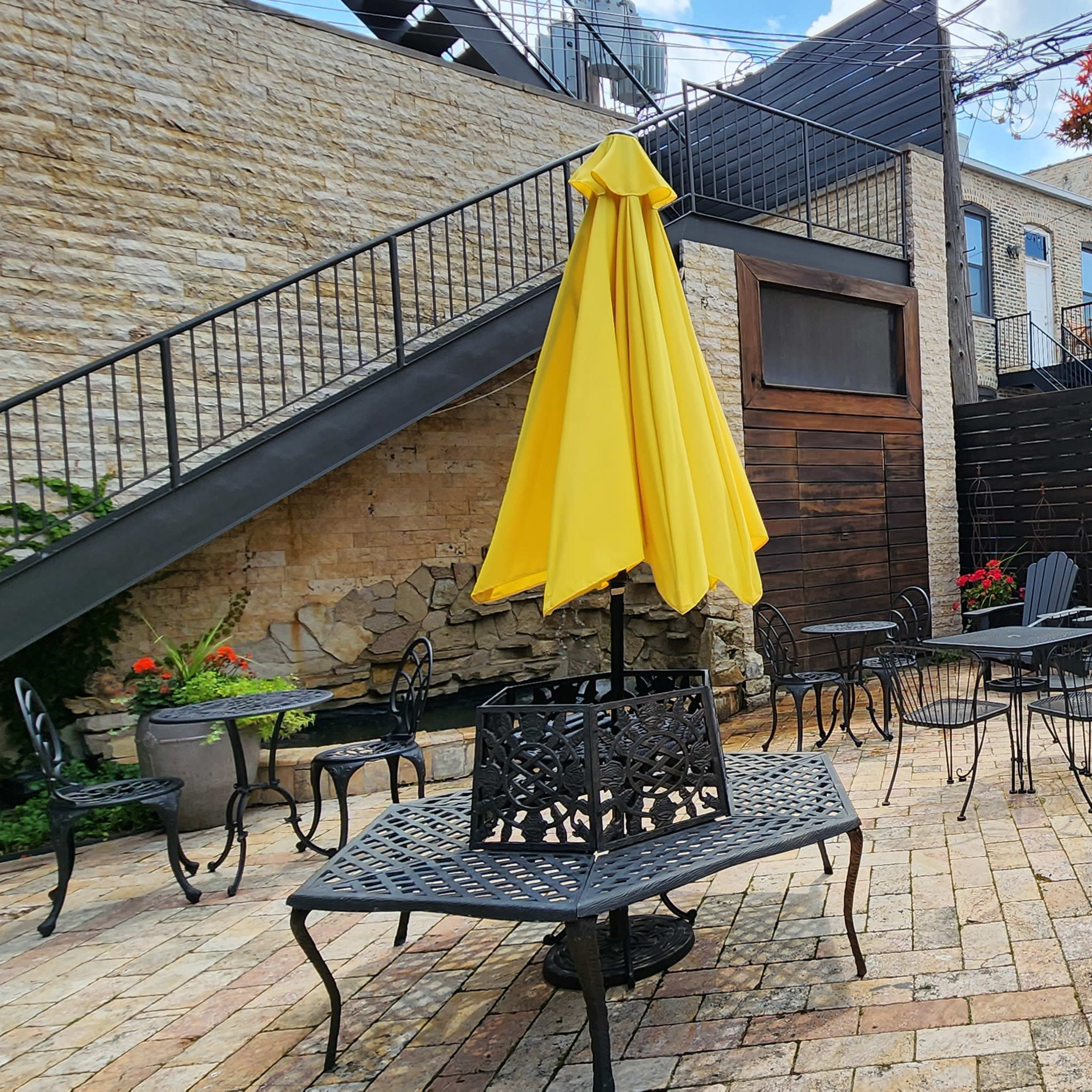 A brick-paved courtyard with several outdoor tables and chairs, a large yellow umbrella, and a small tree surrounded by wooden fences.