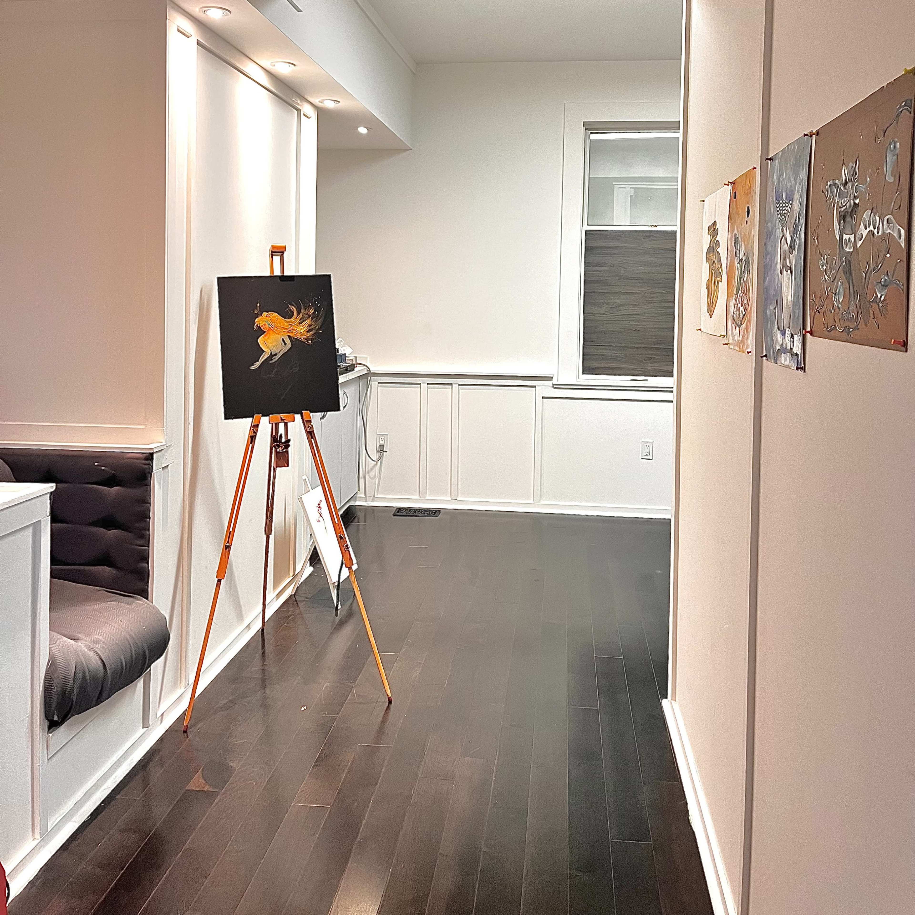 A well-lit room featuring an easel with a painting, hardwood flooring, and a few framed artworks on the wall.