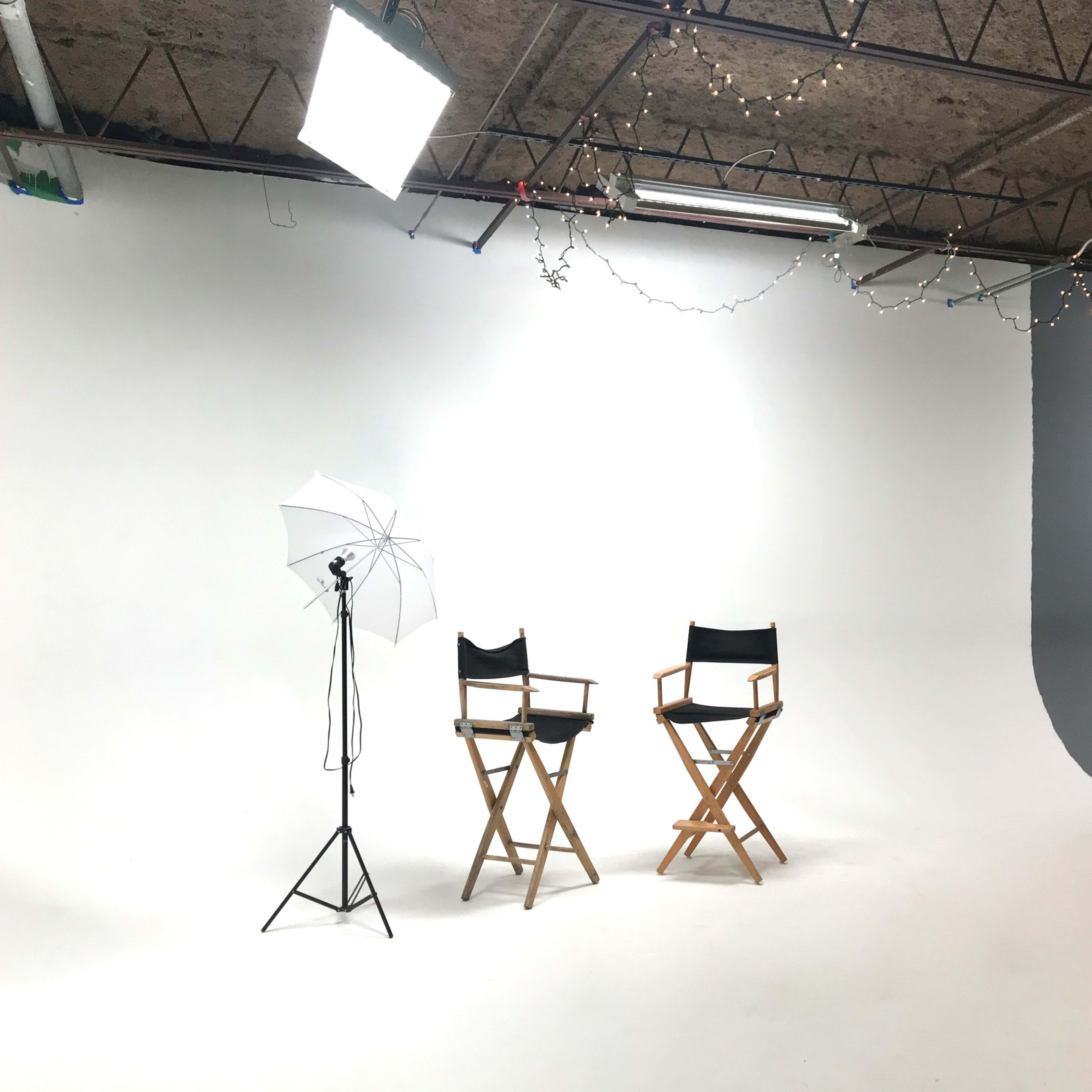 A photography studio features two director's chairs, a lighting setup, and a plain backdrop.